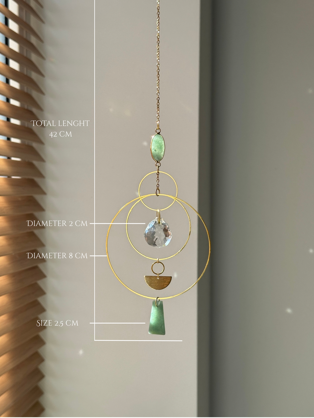 Vertical gold geometric suncatcher with three tiered hoops, a faceted crystal sphere, and two green Aventurine stones casting a galaxy of rainbows on a white wall.