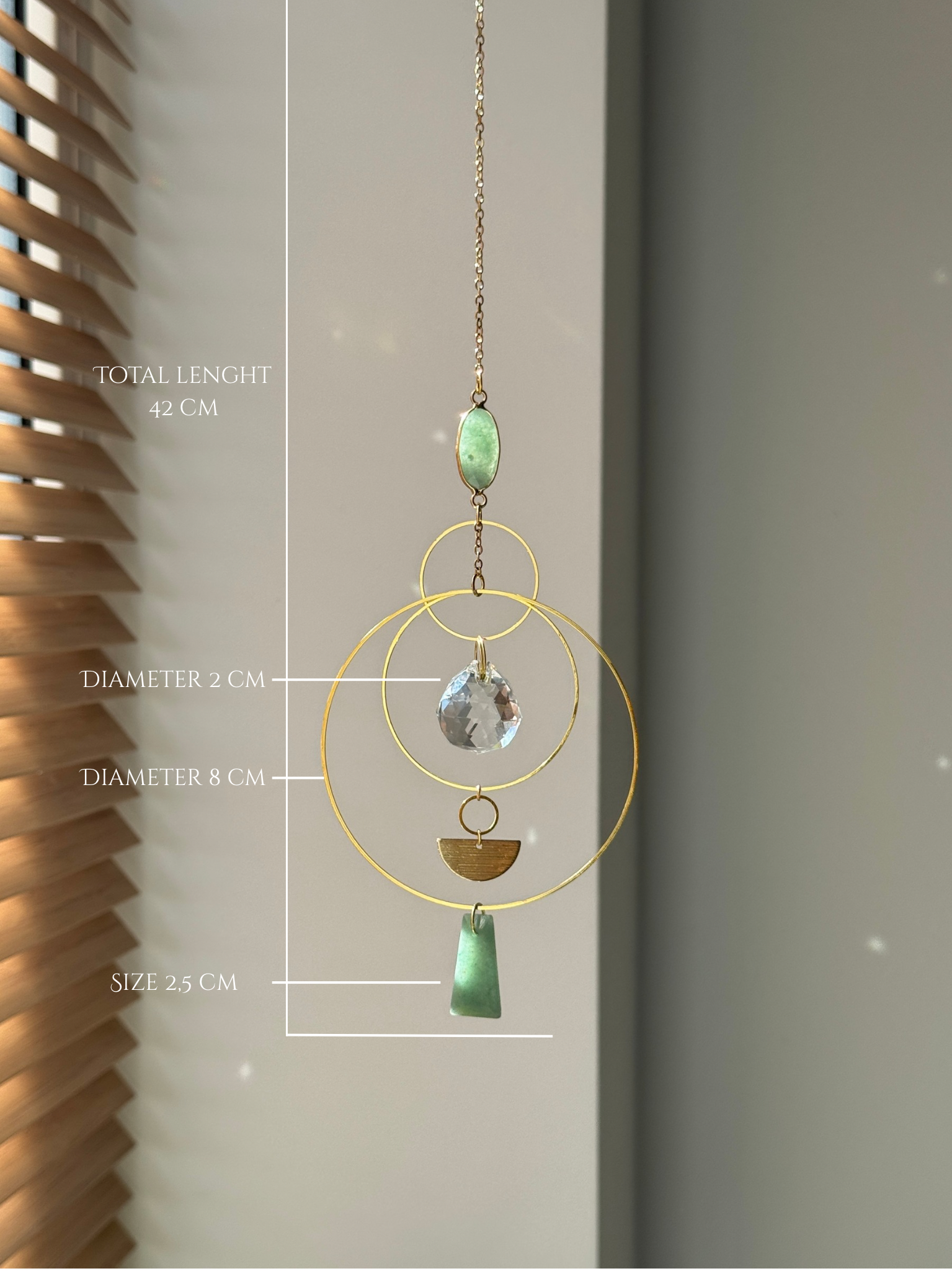 Vertical gold geometric suncatcher with three tiered hoops, a faceted crystal sphere, and two green Aventurine stones casting a galaxy of rainbows on a white wall.