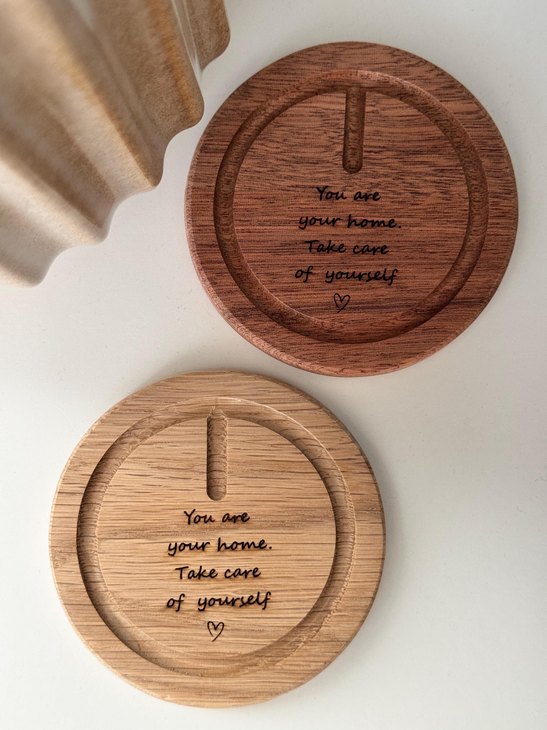 A faceted crystal suncatcher hangs from a rose gold metal hook on a circular wooden base. Sunlight streams through the crystal, scattering vibrant rainbow-colored light reflections across the base and the surrounding white surface. The wooden base is engraved with the message: "You are your home. Take care of yourself." followed by a small hand-drawn heart.