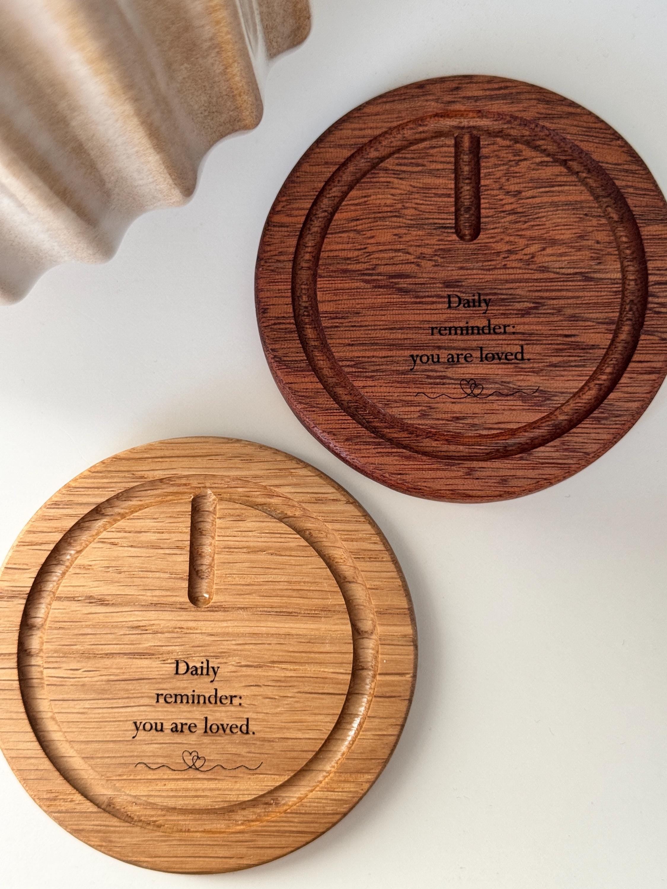 A faceted glass crystal sphere hangs from a gold wire stand on a circular wooden base, which is engraved with the words "Daily reminder: you are loved." The suncatcher is on a white surface next to a wooden window blind. Sunlight streams through the crystal, creating a radiant glow and casting intricate patterns of light and rainbow-colored reflections on the base and surrounding surfaces.