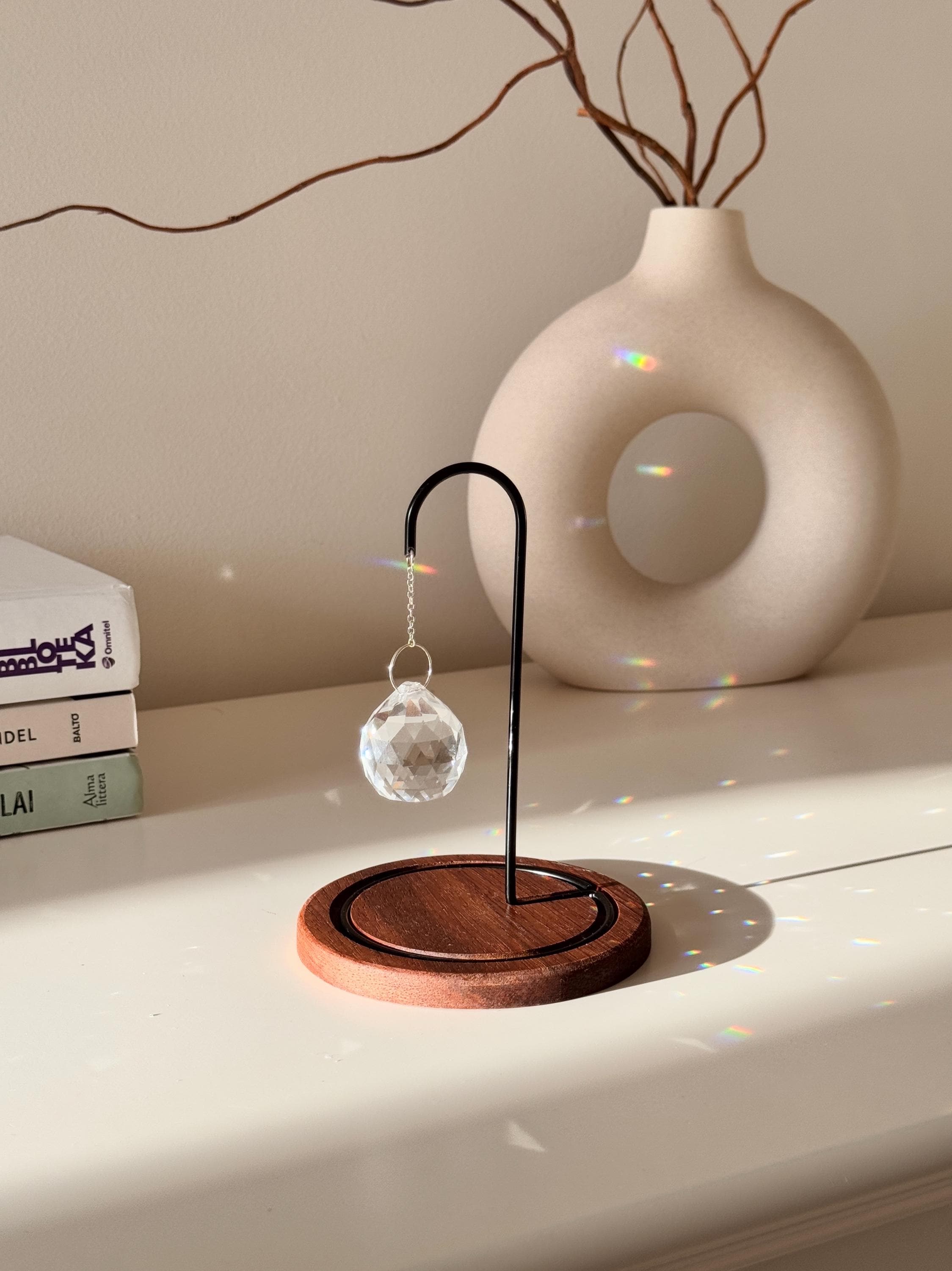 The Aura Hook tabletop suncatcher featuring a faceted crystal prism hanging from a black minimalist stand on a round redwood base, casting rainbow light reflections on a white surface.