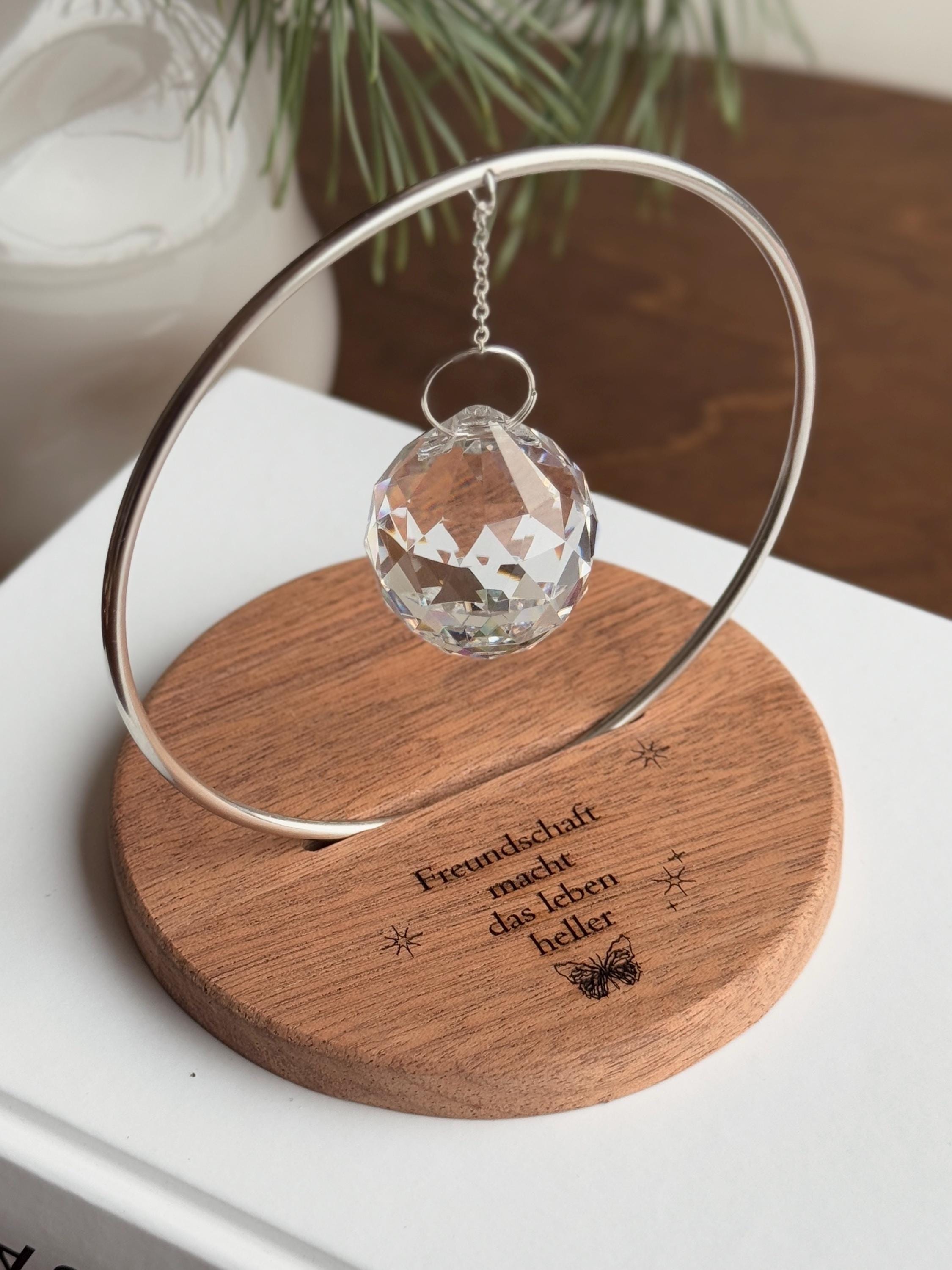 The Circle - A minimalist Sonnenfänger (suncatcher) featuring a high-quality faceted Kristall-Prisma suspended by a delicate gold stainless steel chain within a matching gold ring. The modern tabletop design rests on a solid, dark redwood base (Echtholz-Sockel) and is shown creating vibrant rainbow light reflections (Regenbogen-Effekt) across the background. This elegant Fensterdeko is perfect as a mindful Geschenkidee for home or office.