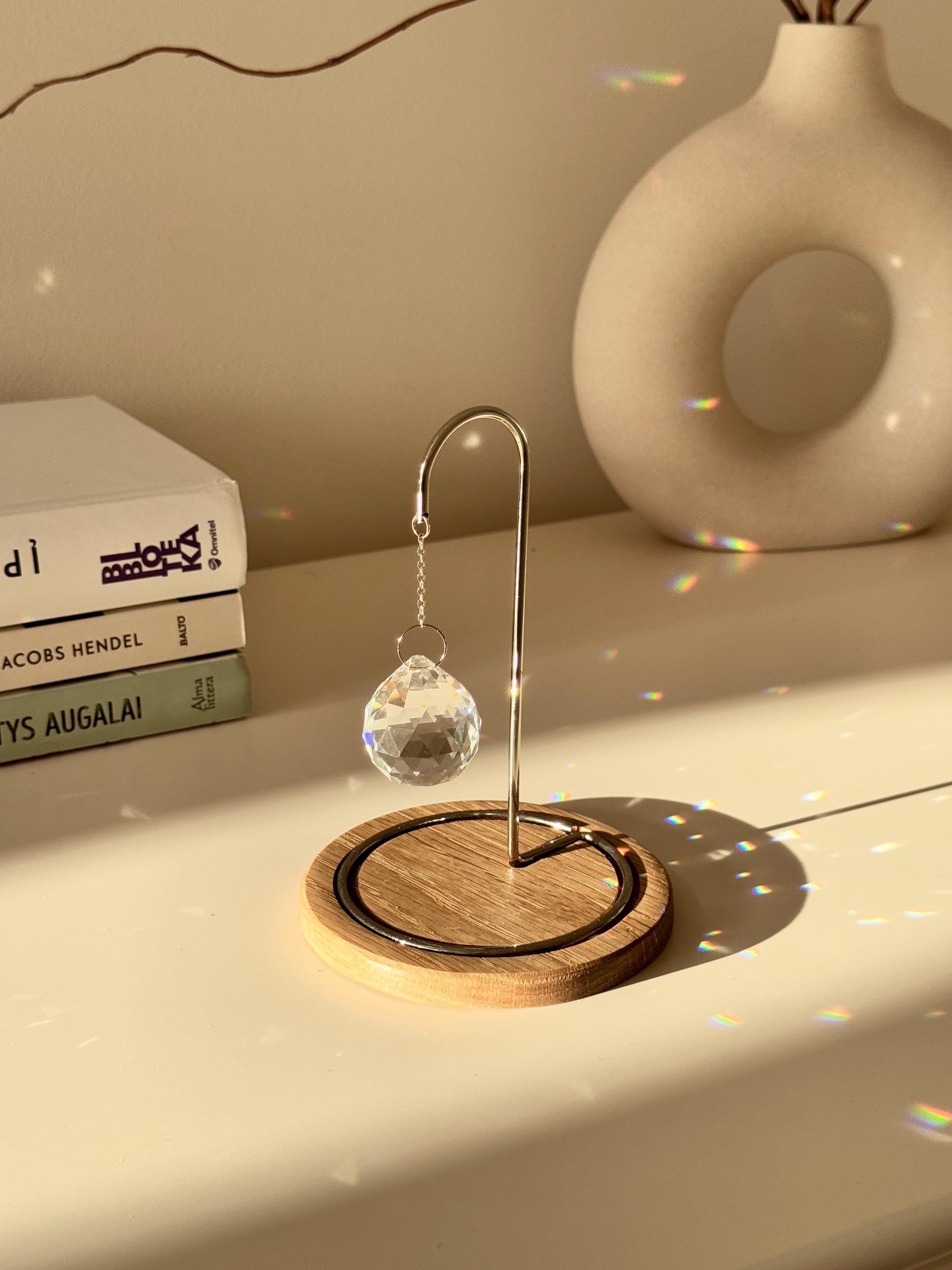 A close-up photograph of a clear, multi-faceted crystal suncatcher prism hanging from an ornate rose gold metal hook on a dark wooden base. The suncatcher casts multiple rainbow-colored refractions of light across the white tabletop, a stack of white books on the left, and a textured off-white ceramic vase in the background. Dried branches are visible emerging from the vase against a light gray wall.
