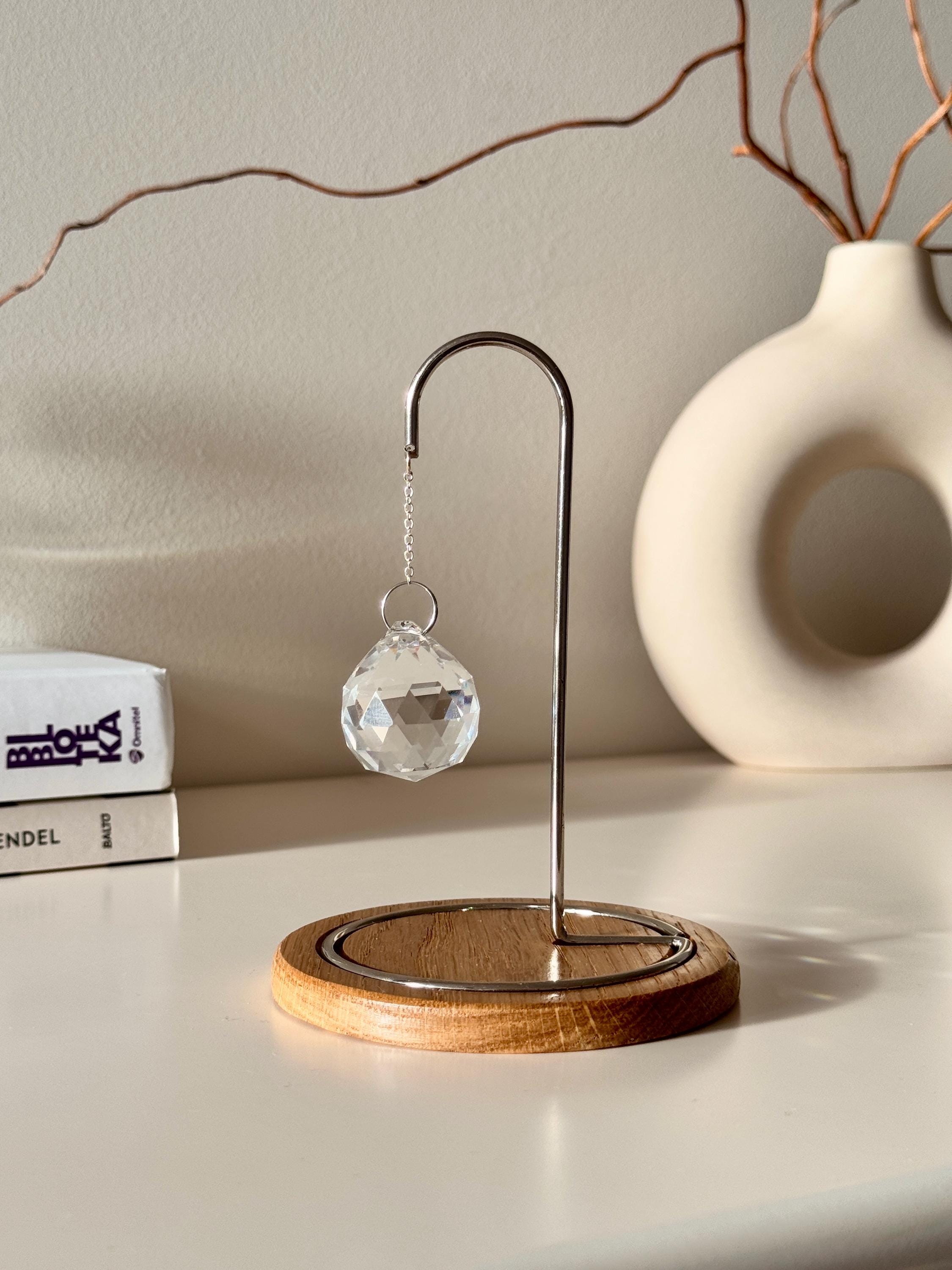 A close-up photograph of a clear, multi-faceted crystal suncatcher prism hanging from an ornate rose gold metal hook on a dark wooden base. The suncatcher casts multiple rainbow-colored refractions of light across the white tabletop, a stack of white books on the left, and a textured off-white ceramic vase in the background. Dried branches are visible emerging from the vase against a light gray wall.
