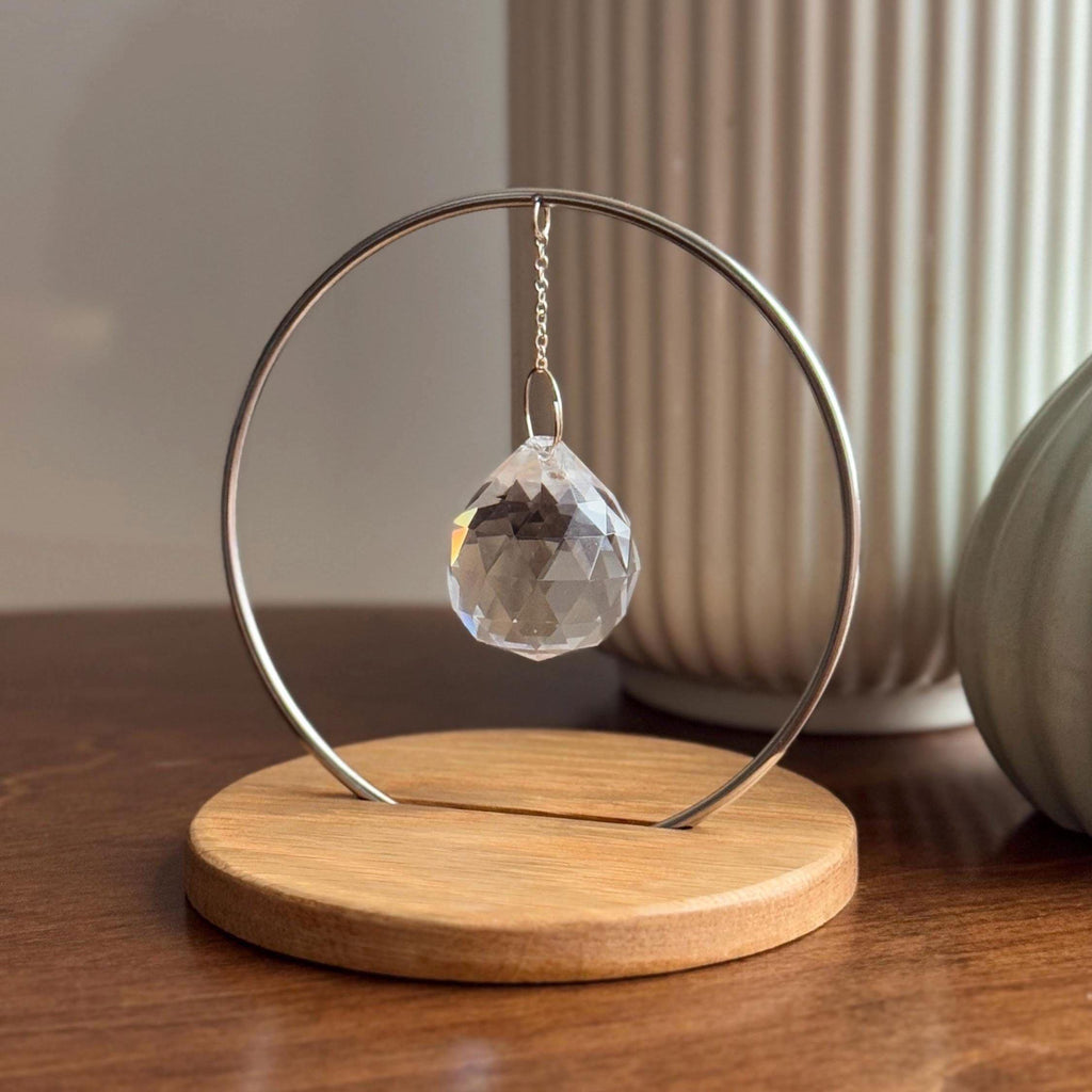 Minimalist "The Circle" tabletop suncatcher featuring a faceted crystal prism hanging inside a stainless steel ring on a round American oak base, casting vibrant rainbows on a white wall.