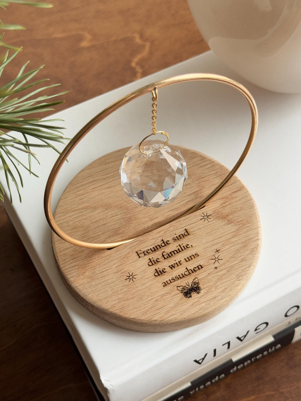 The Circle - A minimalist Sonnenfänger (suncatcher) featuring a high-quality faceted Kristall-Prisma suspended by a delicate gold stainless steel chain within a matching gold ring. The modern tabletop design rests on a solid, dark redwood base (Echtholz-Sockel) and is shown creating vibrant rainbow light reflections (Regenbogen-Effekt) across the background. This elegant Fensterdeko is perfect as a mindful Geschenkidee for home or office.