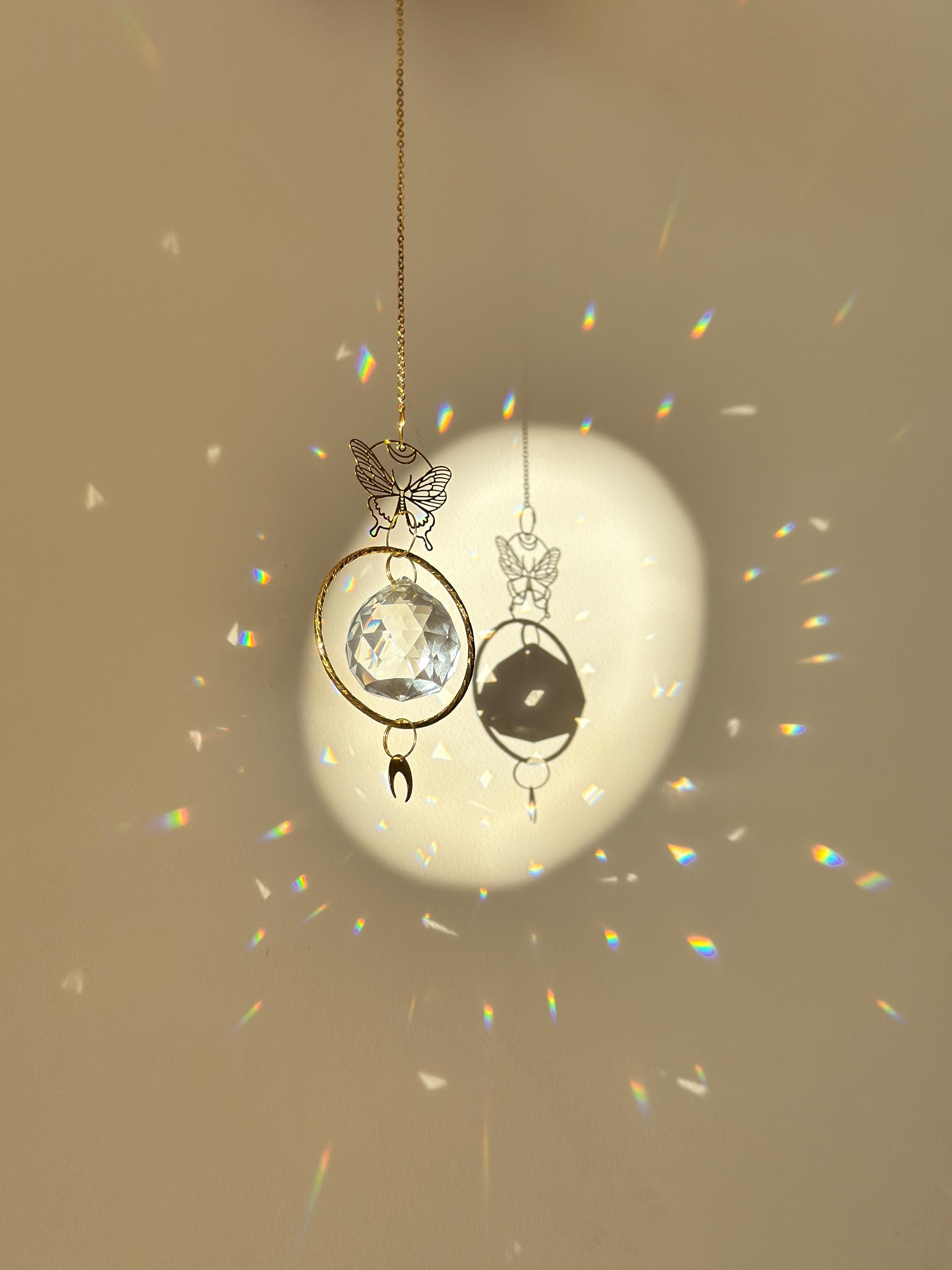 A gold-tone 'Butterfly Effect' Suncatcher features a delicate, etched butterfly charm at the top, followed by a large, circular brass frame housing a premium 40mm faceted crystal prism. A small, downward-facing crescent moon dangles at the very bottom. Direct sunlight illuminates the glass, scattering a magical "Meer von Regenbögen" (sea of rainbows) and "Sternenstaub" (stardust) across the warm, neutral wall. The suncatcher casts a sharp, elegant shadow of the butterfly and moon motifs, creating a serene s
