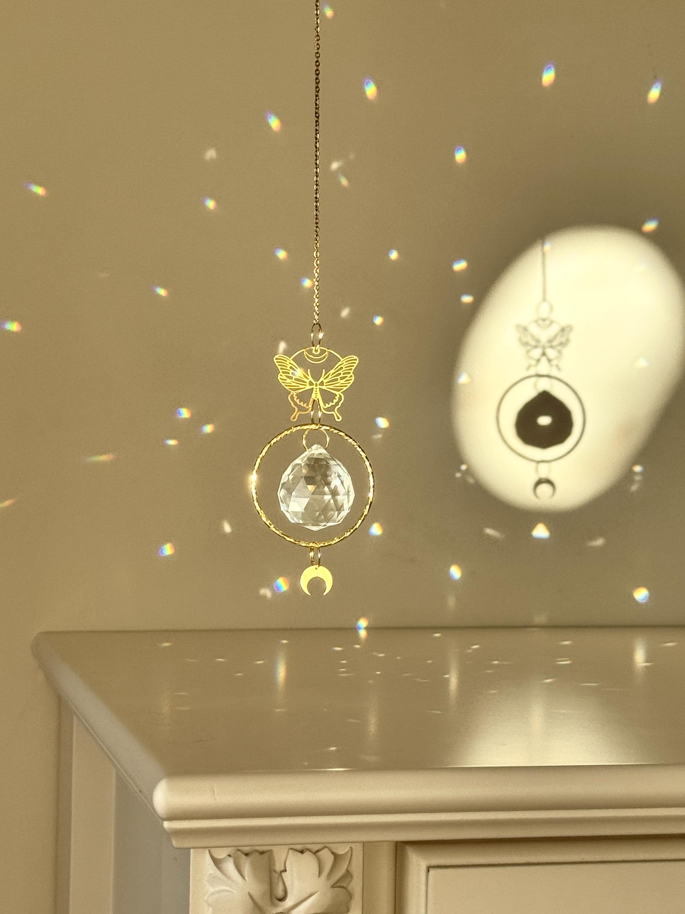 A gold-tone 'Butterfly Effect' Suncatcher features a delicate, etched butterfly charm at the top, followed by a large, circular brass frame housing a premium 40mm faceted crystal prism. A small, downward-facing crescent moon dangles at the very bottom. Direct sunlight illuminates the glass, scattering a magical "Meer von Regenbögen" (sea of rainbows) and "Sternenstaub" (stardust) across the warm, neutral wall. The suncatcher casts a sharp, elegant shadow of the butterfly and moon motifs, creating a serene s