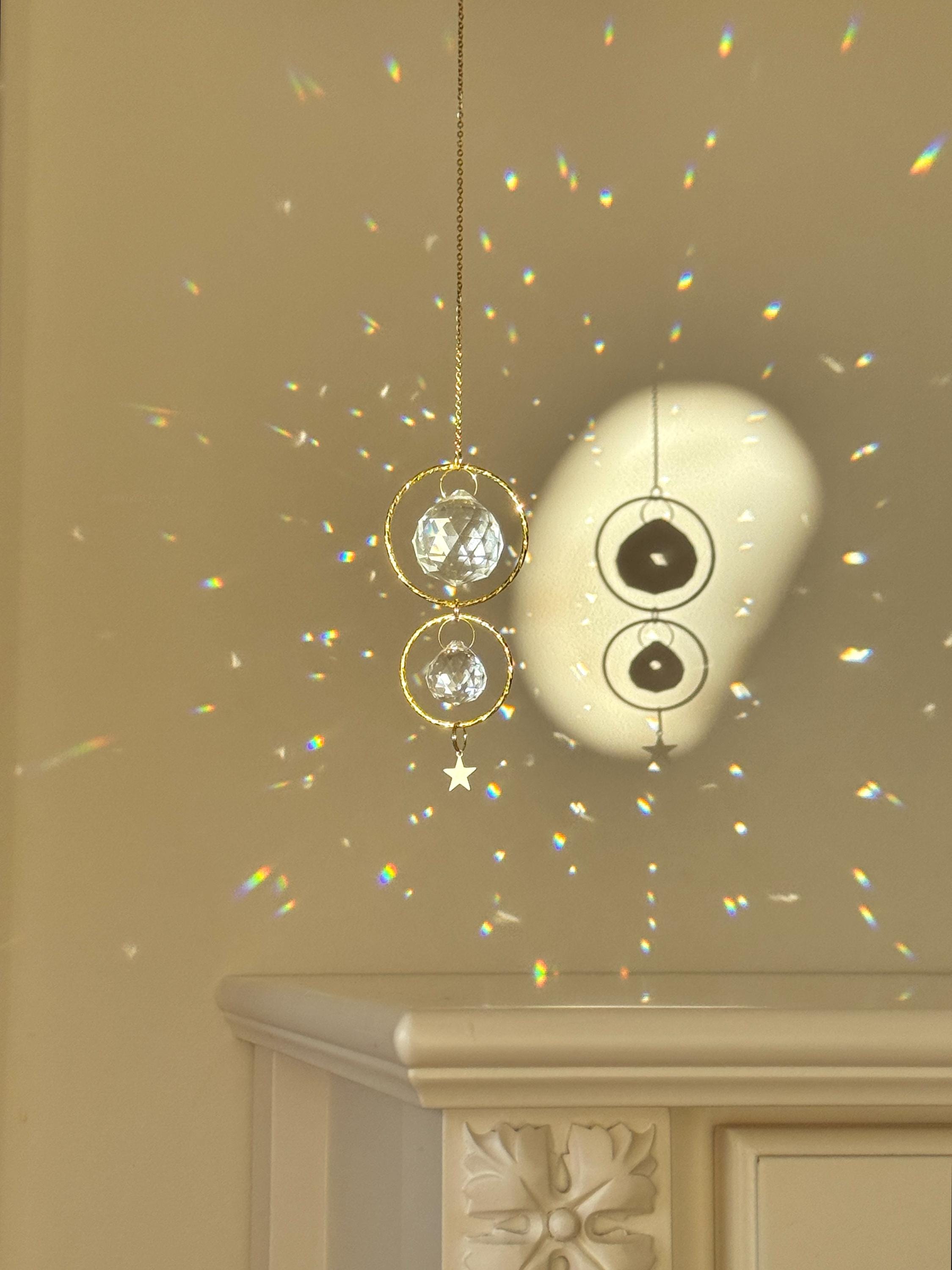 A gold-tone 'Double Harmony' Suncatcher featuring two geometric frames suspended on a delicate chain. The frames house a duo of faceted crystal prisms (20mm and 40mm) that capture the light to create a vibrant "Meer von Regenbögen" (sea of rainbows) on the surrounding walls. The design exudes Gemütlichkeit (coziness), casting elegant shadows and "Sternenstaub" (stardust) reflections across a modern, sunlit interior.