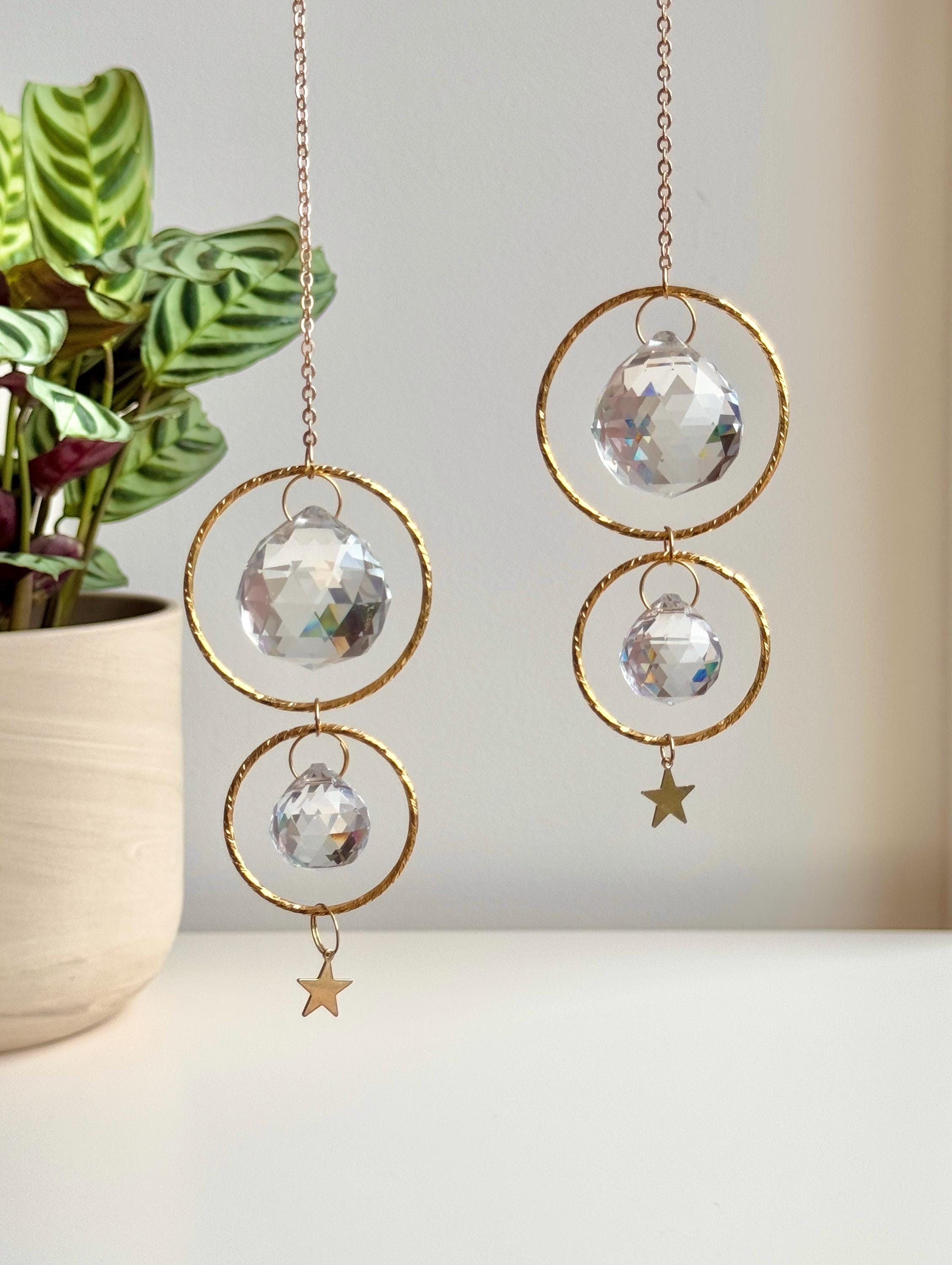 A gold-tone 'Double Harmony' Suncatcher featuring two geometric frames suspended on a delicate chain. The frames house a duo of faceted crystal prisms (20mm and 40mm) that capture the light to create a vibrant "Meer von Regenbögen" (sea of rainbows) on the surrounding walls. The design exudes Gemütlichkeit (coziness), casting elegant shadows and "Sternenstaub" (stardust) reflections across a modern, sunlit interior.