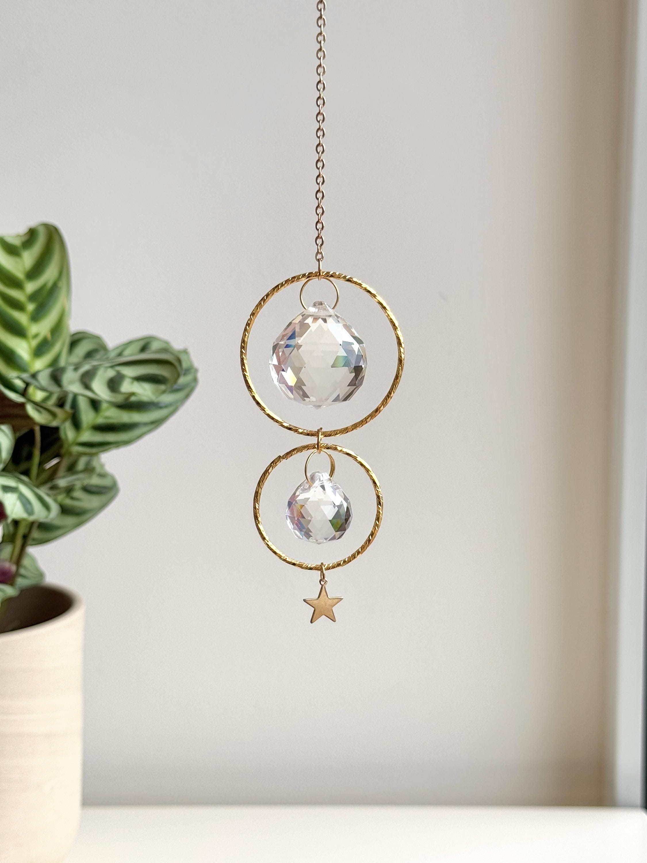 A gold-tone 'Double Harmony' Suncatcher featuring two geometric frames suspended on a delicate chain. The frames house a duo of faceted crystal prisms (20mm and 40mm) that capture the light to create a vibrant "Meer von Regenbögen" (sea of rainbows) on the surrounding walls. The design exudes Gemütlichkeit (coziness), casting elegant shadows and "Sternenstaub" (stardust) reflections across a modern, sunlit interior.