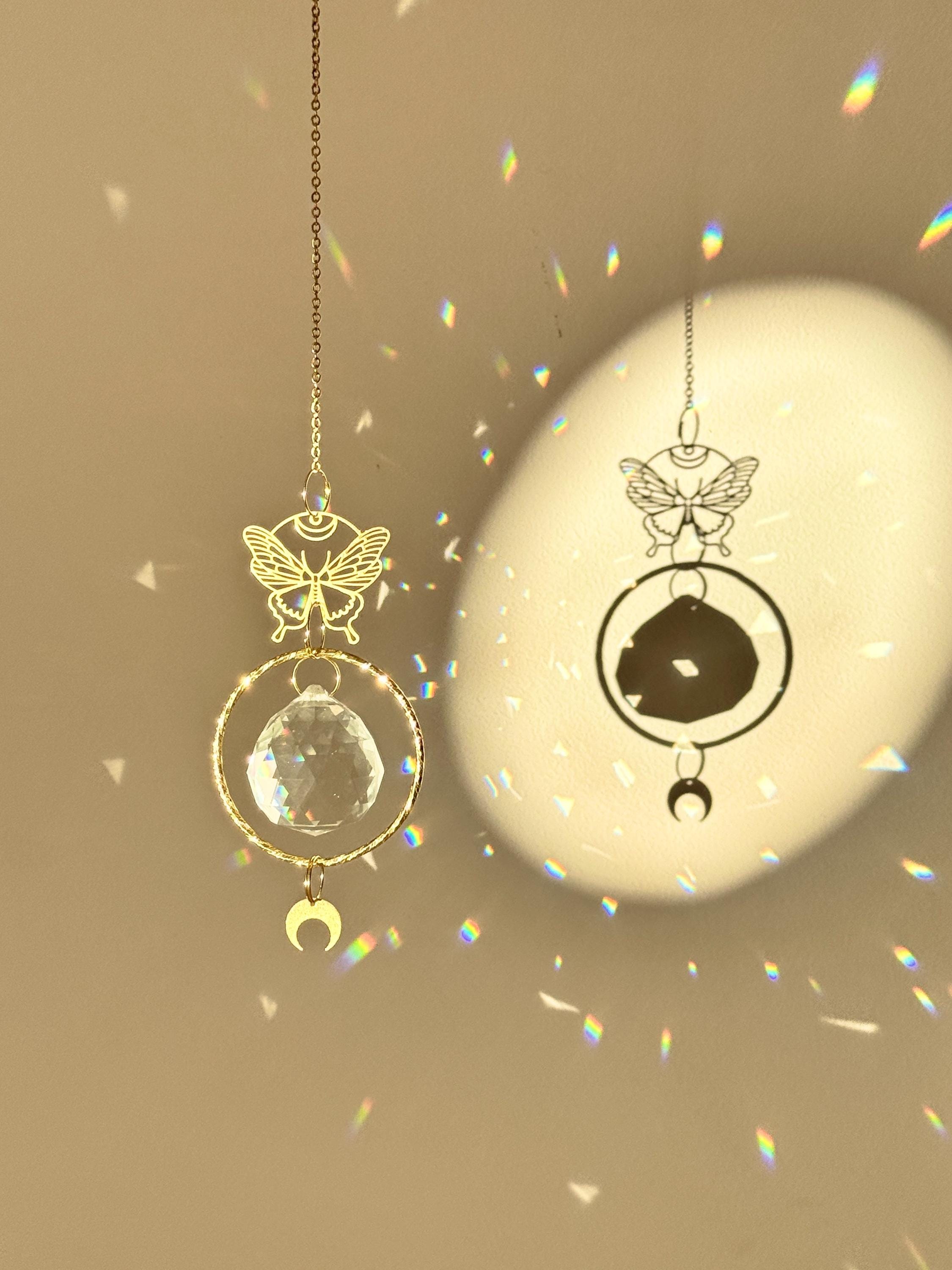A gold-tone 'Butterfly Effect' Suncatcher features a delicate, etched butterfly charm at the top, followed by a large, circular brass frame housing a premium 40mm faceted crystal prism. A small, downward-facing crescent moon dangles at the very bottom. Direct sunlight illuminates the glass, scattering a magical "Meer von Regenbögen" (sea of rainbows) and "Sternenstaub" (stardust) across the warm, neutral wall. The suncatcher casts a sharp, elegant shadow of the butterfly and moon motifs, creating a serene s