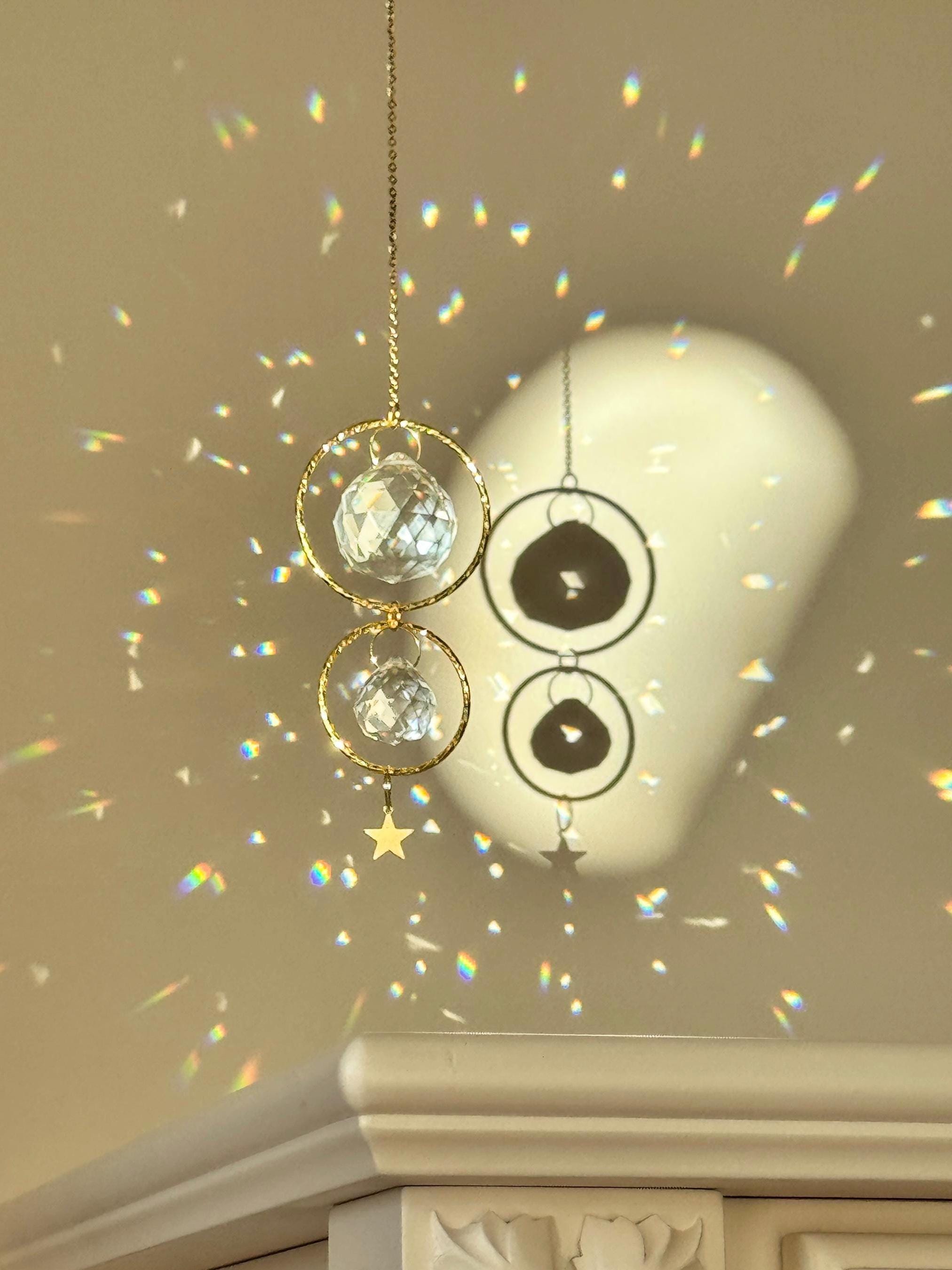 A gold-tone 'Double Harmony' Suncatcher featuring two geometric frames suspended on a delicate chain. The frames house a duo of faceted crystal prisms (20mm and 40mm) that capture the light to create a vibrant "Meer von Regenbögen" (sea of rainbows) on the surrounding walls. The design exudes Gemütlichkeit (coziness), casting elegant shadows and "Sternenstaub" (stardust) reflections across a modern, sunlit interior.