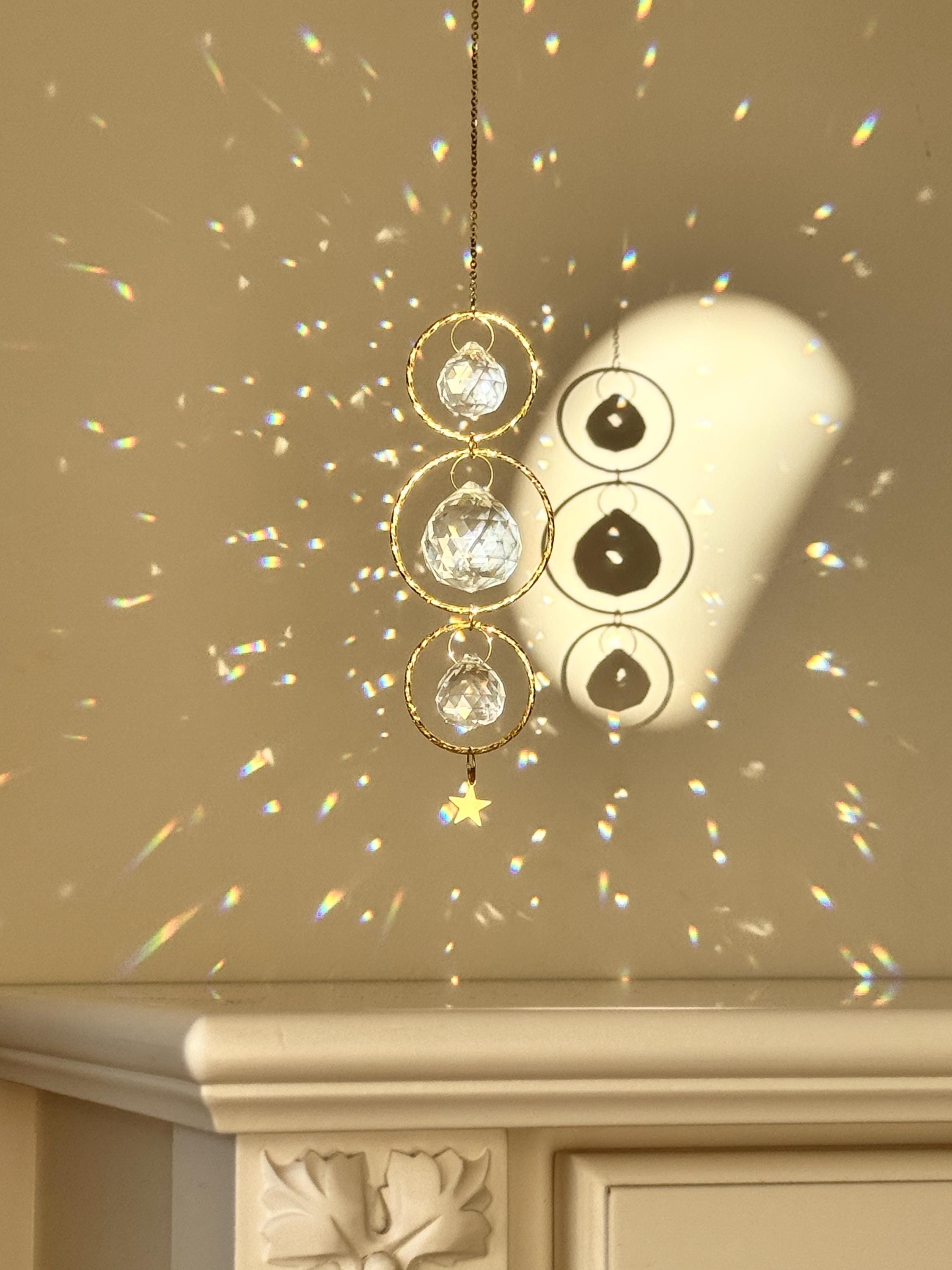 A close-up of the 'Triple Harmony' Suncatcher showing three interlocking gold hoops, each suspending a high-quality faceted glass prism. The central crystal is larger (40mm) than the top and bottom ones (20mm), creating a balanced, tiered effect. The sunlight transforms the room into a canvas of shimmering spectral light and "Sternenstaub" (stardust) reflections. A sharp, rhythmic shadow of the celestial design is cast on the wall behind, highlighting the boho-chic aesthetic.