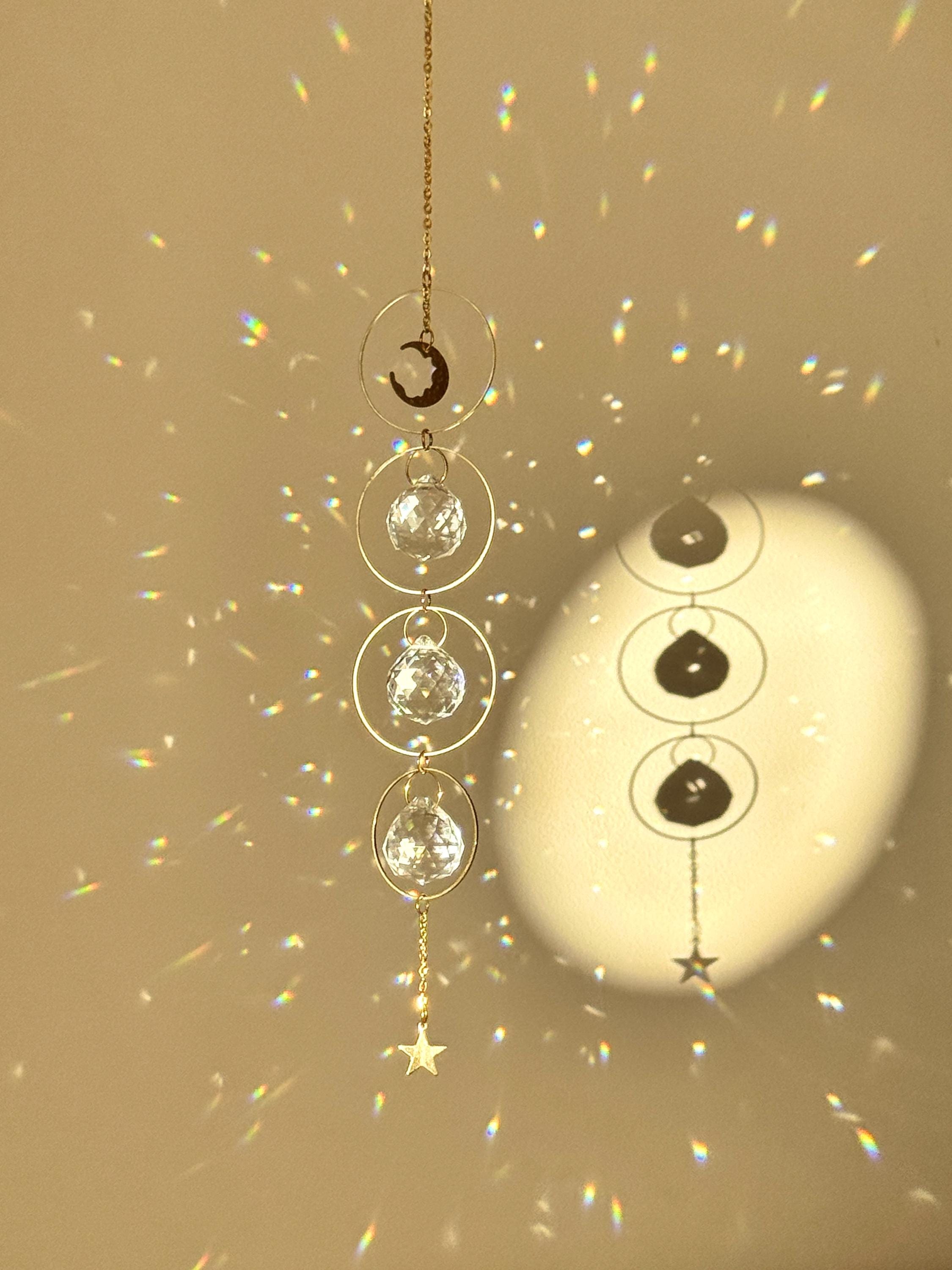 A gold-tone 'Serenity' celestial suncatcher hangs against a warm, neutral wall, featuring three circular brass hoops. The top hoop holds a delicate crescent moon, while the bottom three each frame a 20mm faceted crystal prism that ends in a small gold star. Bright sunlight creates a magical "Meer von Regenbögen" (sea of rainbows) and shimmering light speckles across the surface. To the right, a reed diffuser and stacked books create a sense of Gemütlichkeit (coziness), while the suncatcher casts a soft, rhy