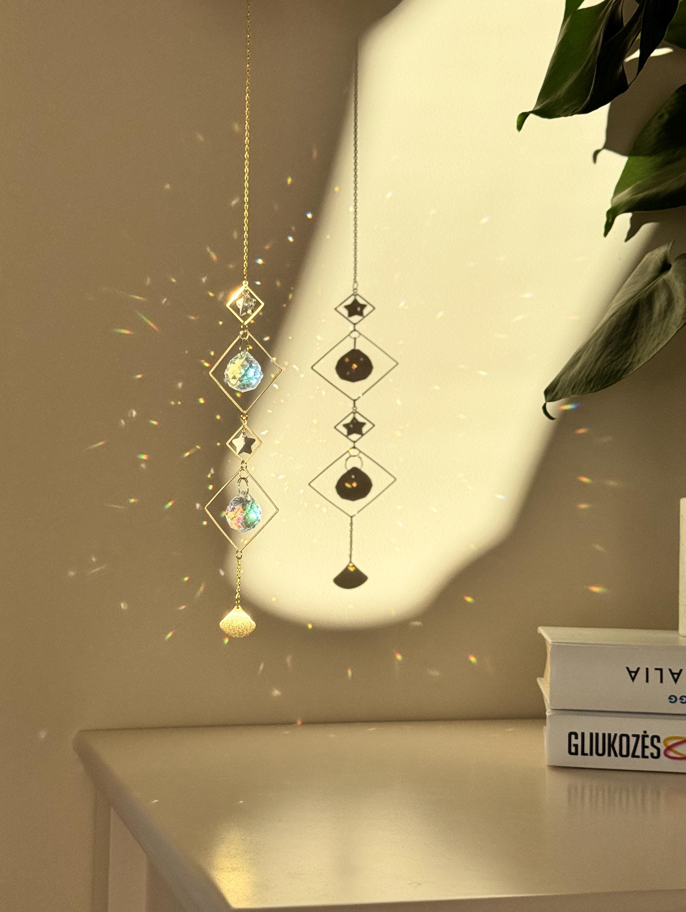 A vertical gold-tone geometric suncatcher hangs elegantly against a cream-colored wall, casting a dazzling "Meer von Regenbögen" (sea of rainbows) across the room. The piece features alternating small and large diamond-shaped brass frames, with two faceted 2cm crystal prisms suspended in the center. Sunlight passes through the glass, creating vibrant spectral flares that dance like "Sternenstaub" (stardust). To the right, a lush green Monstera leaf and a stack of books add a modern, boho-chic vibe. The over
