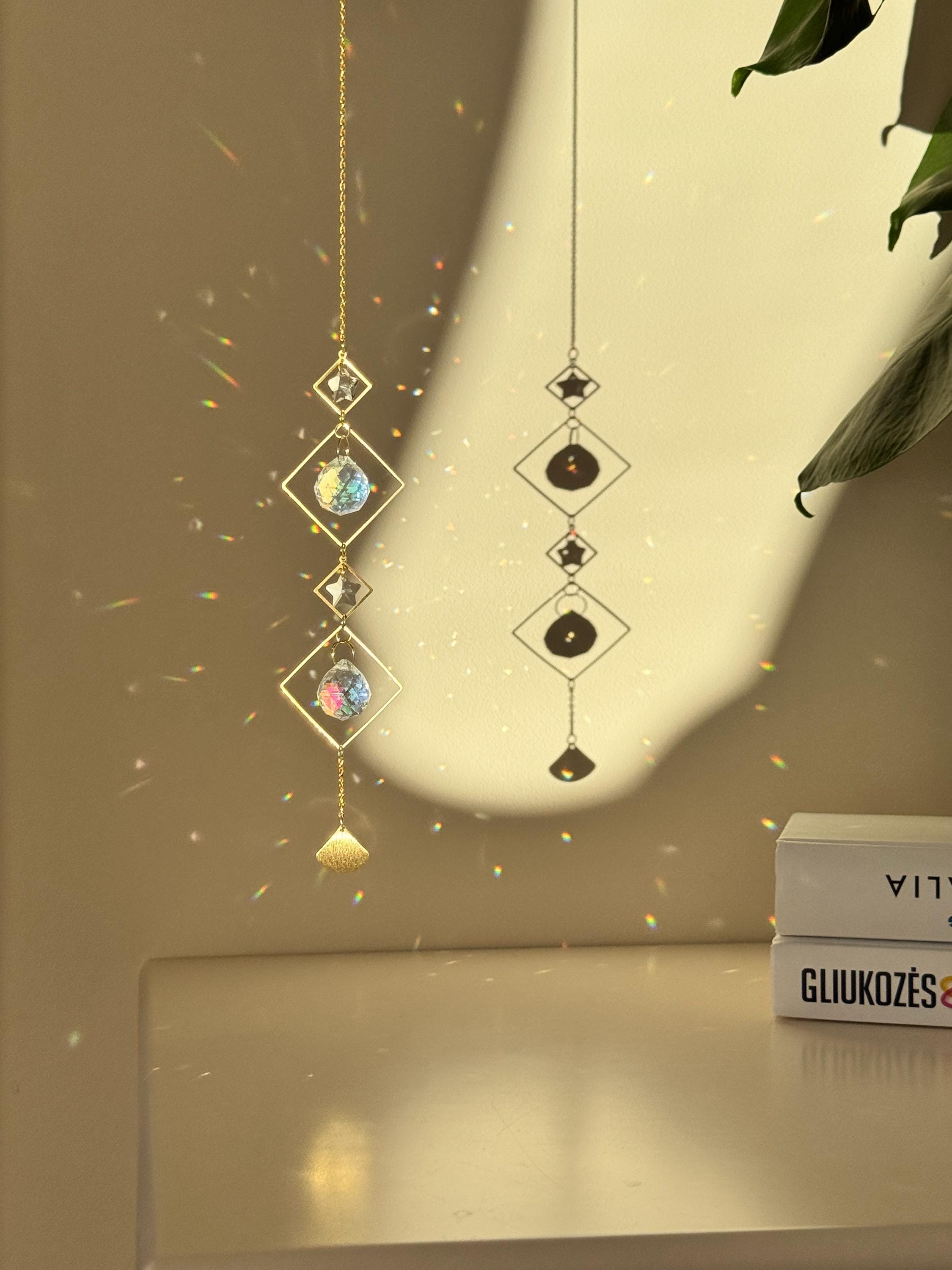 A vertical gold-tone geometric suncatcher hangs elegantly against a cream-colored wall, casting a dazzling "Meer von Regenbögen" (sea of rainbows) across the room. The piece features alternating small and large diamond-shaped brass frames, with two faceted 2cm crystal prisms suspended in the center. Sunlight passes through the glass, creating vibrant spectral flares that dance like "Sternenstaub" (stardust). To the right, a lush green Monstera leaf and a stack of books add a modern, boho-chic vibe. The over