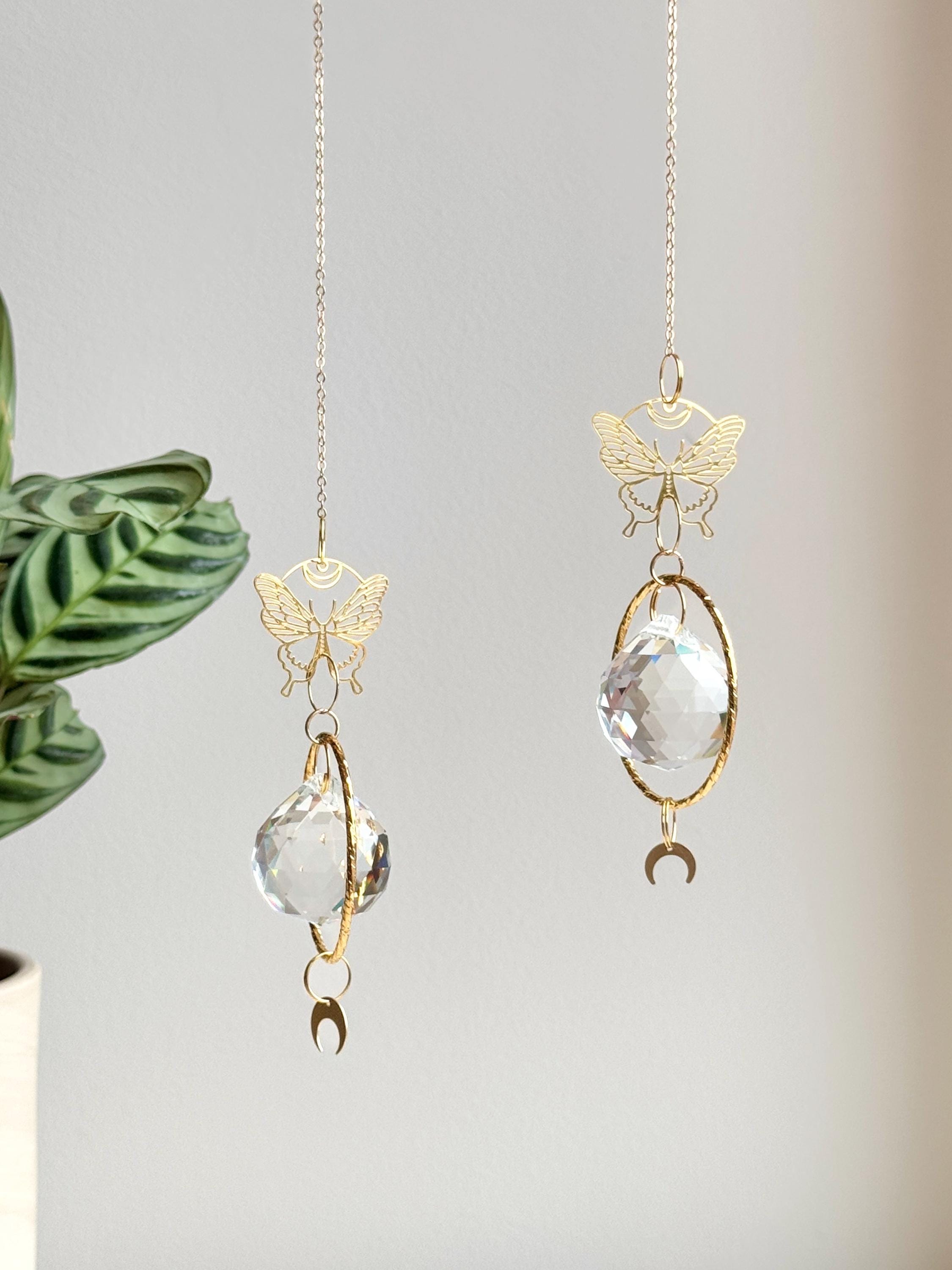 A gold-tone 'Butterfly Effect' Suncatcher features a delicate, etched butterfly charm at the top, followed by a large, circular brass frame housing a premium 40mm faceted crystal prism. A small, downward-facing crescent moon dangles at the very bottom. Direct sunlight illuminates the glass, scattering a magical "Meer von Regenbögen" (sea of rainbows) and "Sternenstaub" (stardust) across the warm, neutral wall. The suncatcher casts a sharp, elegant shadow of the butterfly and moon motifs, creating a serene s