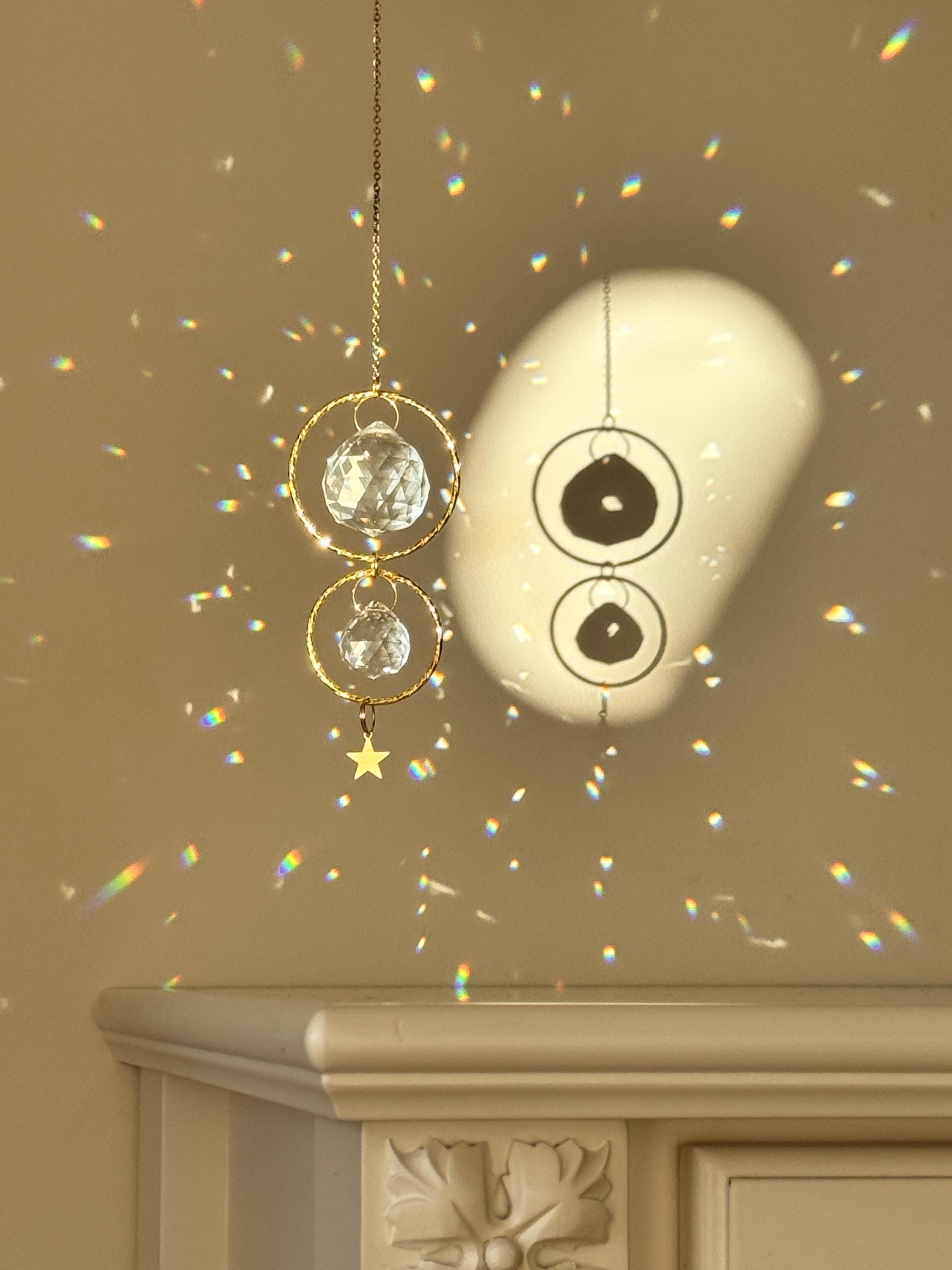 A gold-tone 'Double Harmony' Suncatcher featuring two geometric frames suspended on a delicate chain. The frames house a duo of faceted crystal prisms (20mm and 40mm) that capture the light to create a vibrant "Meer von Regenbögen" (sea of rainbows) on the surrounding walls. The design exudes Gemütlichkeit (coziness), casting elegant shadows and "Sternenstaub" (stardust) reflections across a modern, sunlit interior.