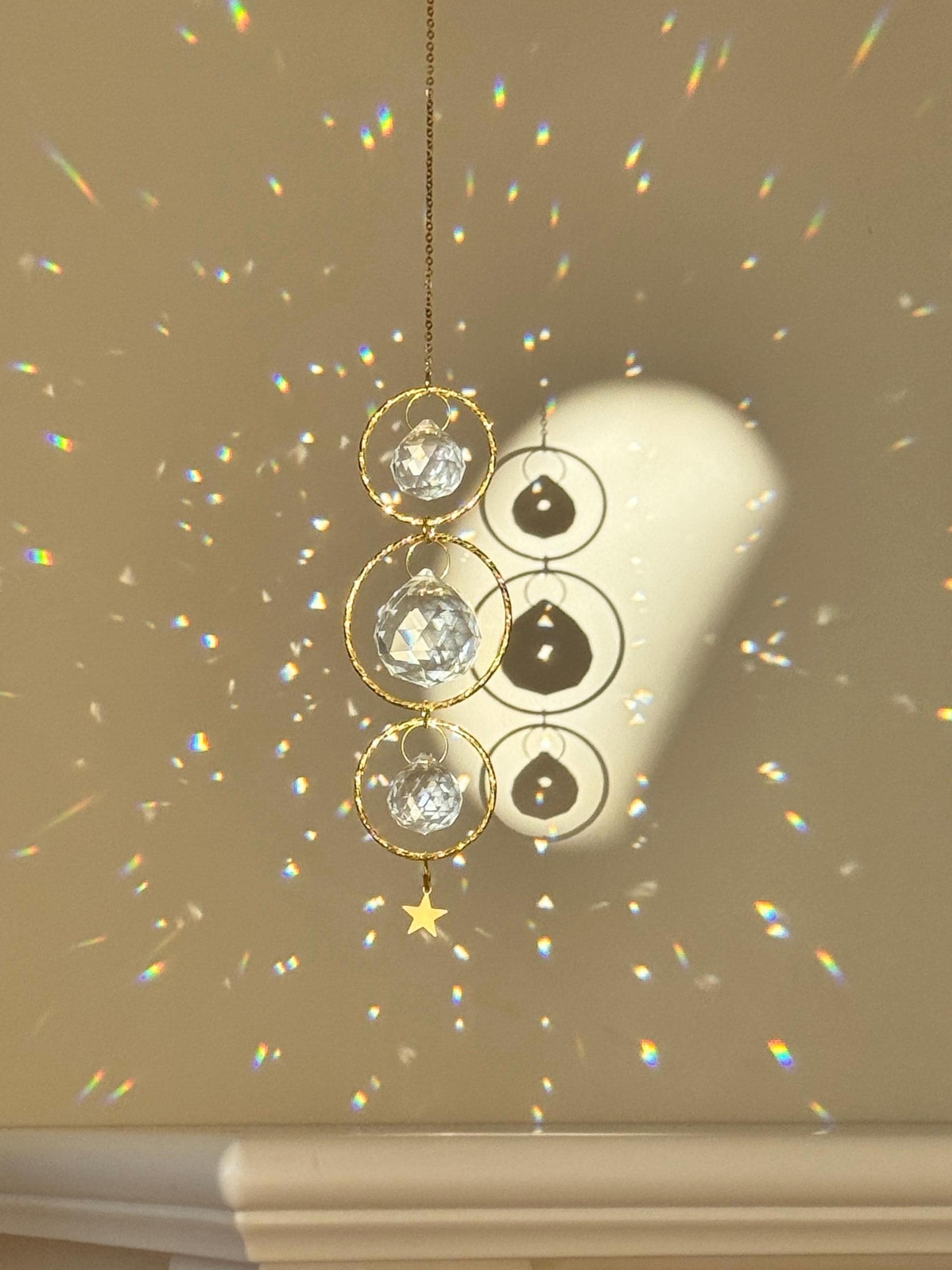 A close-up of the 'Triple Harmony' Suncatcher showing three interlocking gold hoops, each suspending a high-quality faceted glass prism. The central crystal is larger (40mm) than the top and bottom ones (20mm), creating a balanced, tiered effect. The sunlight transforms the room into a canvas of shimmering spectral light and "Sternenstaub" (stardust) reflections. A sharp, rhythmic shadow of the celestial design is cast on the wall behind, highlighting the boho-chic aesthetic.