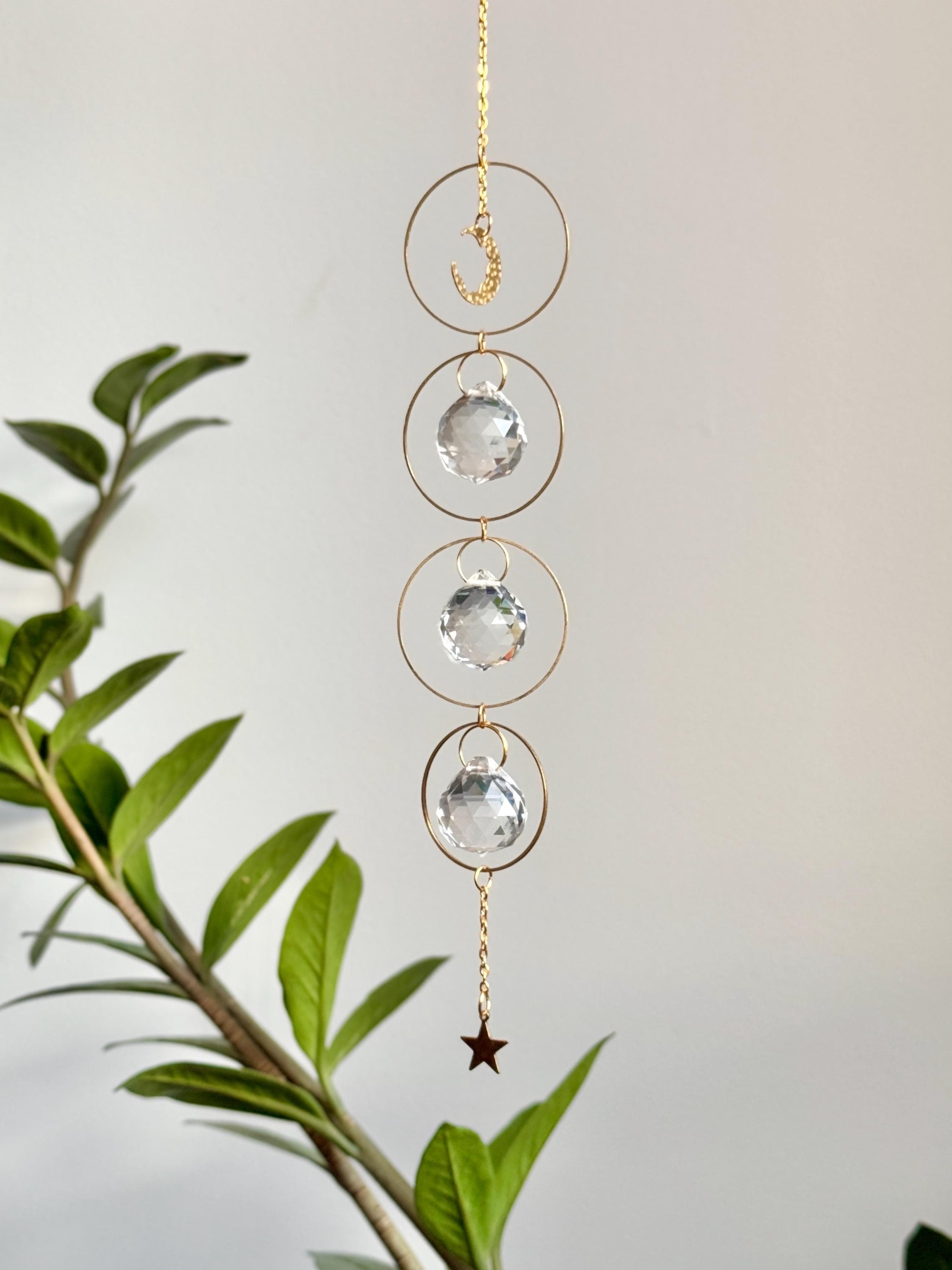 A gold-tone 'Serenity' celestial suncatcher hangs against a warm, neutral wall, featuring three circular brass hoops. The top hoop holds a delicate crescent moon, while the bottom three each frame a 20mm faceted crystal prism that ends in a small gold star. Bright sunlight creates a magical "Meer von Regenbögen" (sea of rainbows) and shimmering light speckles across the surface. To the right, a reed diffuser and stacked books create a sense of Gemütlichkeit (coziness), while the suncatcher casts a soft, rhy