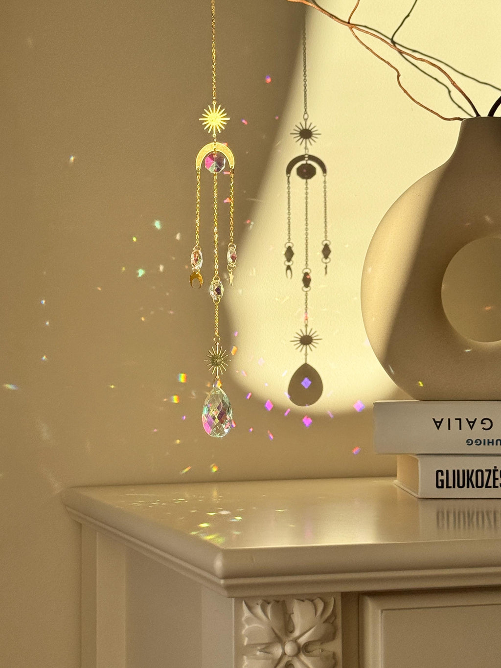 The 'Cosmic Balance' suncatcher, 47cm long, featuring a gold celestial geometric frame and a large 4cm crystal prism, hanging in a bright window and casting wide rainbow patterns.