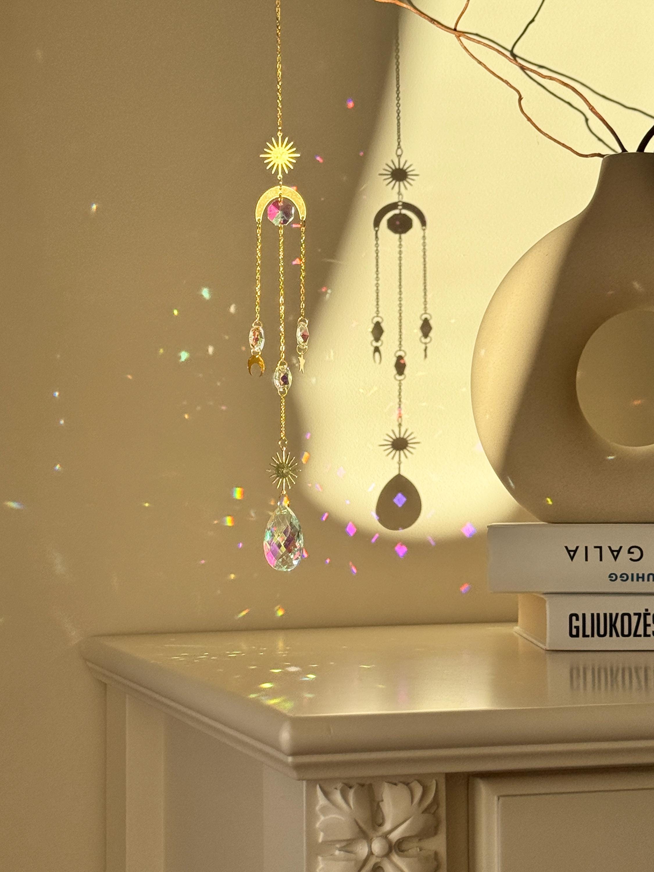 The 'Cosmic Balance' suncatcher, 47cm long, featuring a gold celestial geometric frame and a large 4cm crystal prism, hanging in a bright window and casting wide rainbow patterns.