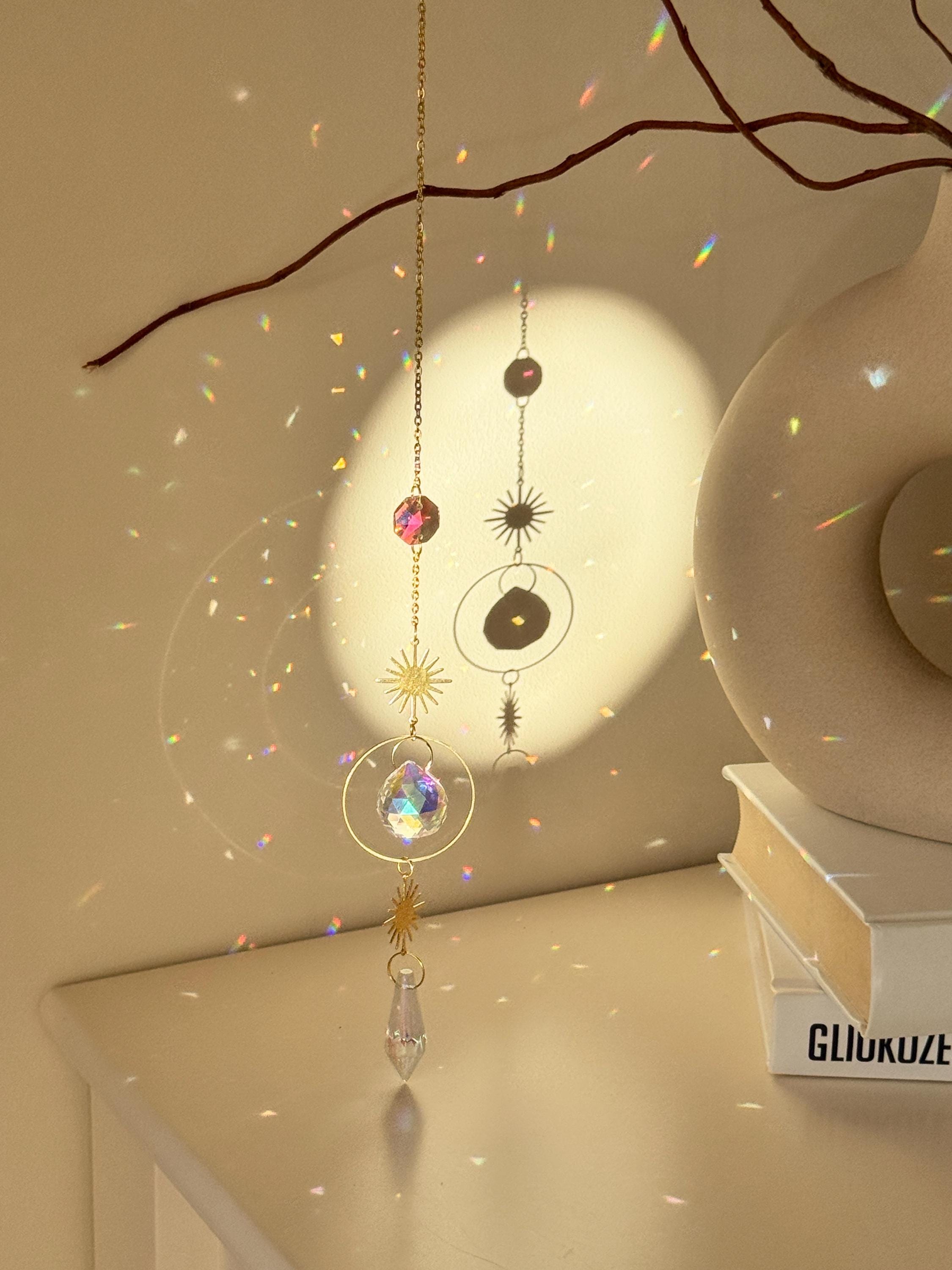 Celestial 'Sunny Days' suncatcher with gold sun charms and two crystal prisms hanging against a warm-toned wall, casting small rainbow light spots across the room.