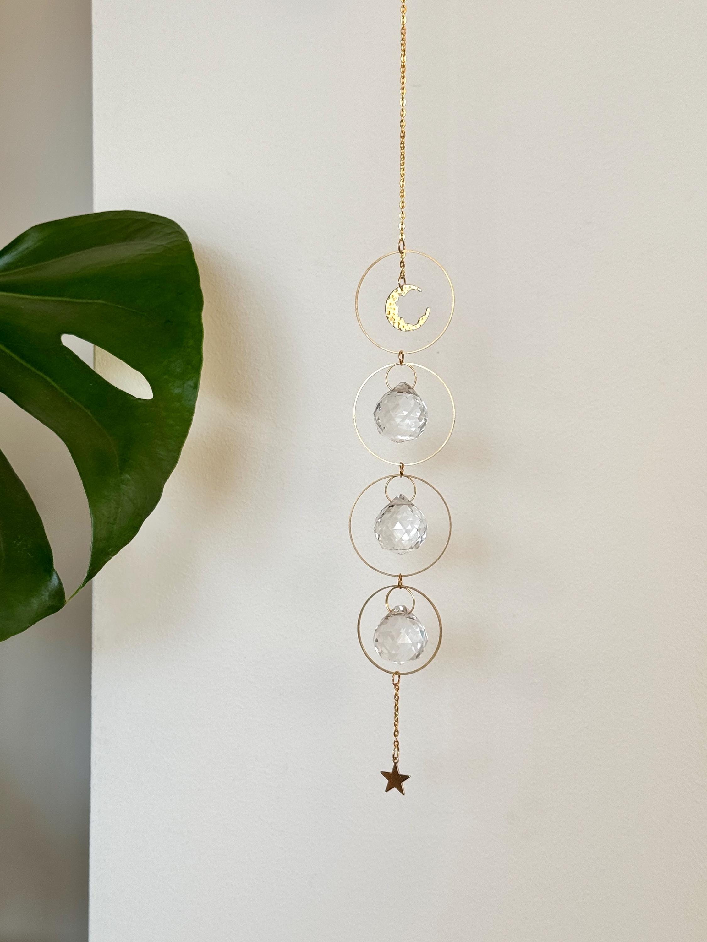 A gold-tone 'Serenity' celestial suncatcher hangs against a warm, neutral wall, featuring three circular brass hoops. The top hoop holds a delicate crescent moon, while the bottom three each frame a 20mm faceted crystal prism that ends in a small gold star. Bright sunlight creates a magical "Meer von Regenbögen" (sea of rainbows) and shimmering light speckles across the surface. To the right, a reed diffuser and stacked books create a sense of Gemütlichkeit (coziness), while the suncatcher casts a soft, rhy