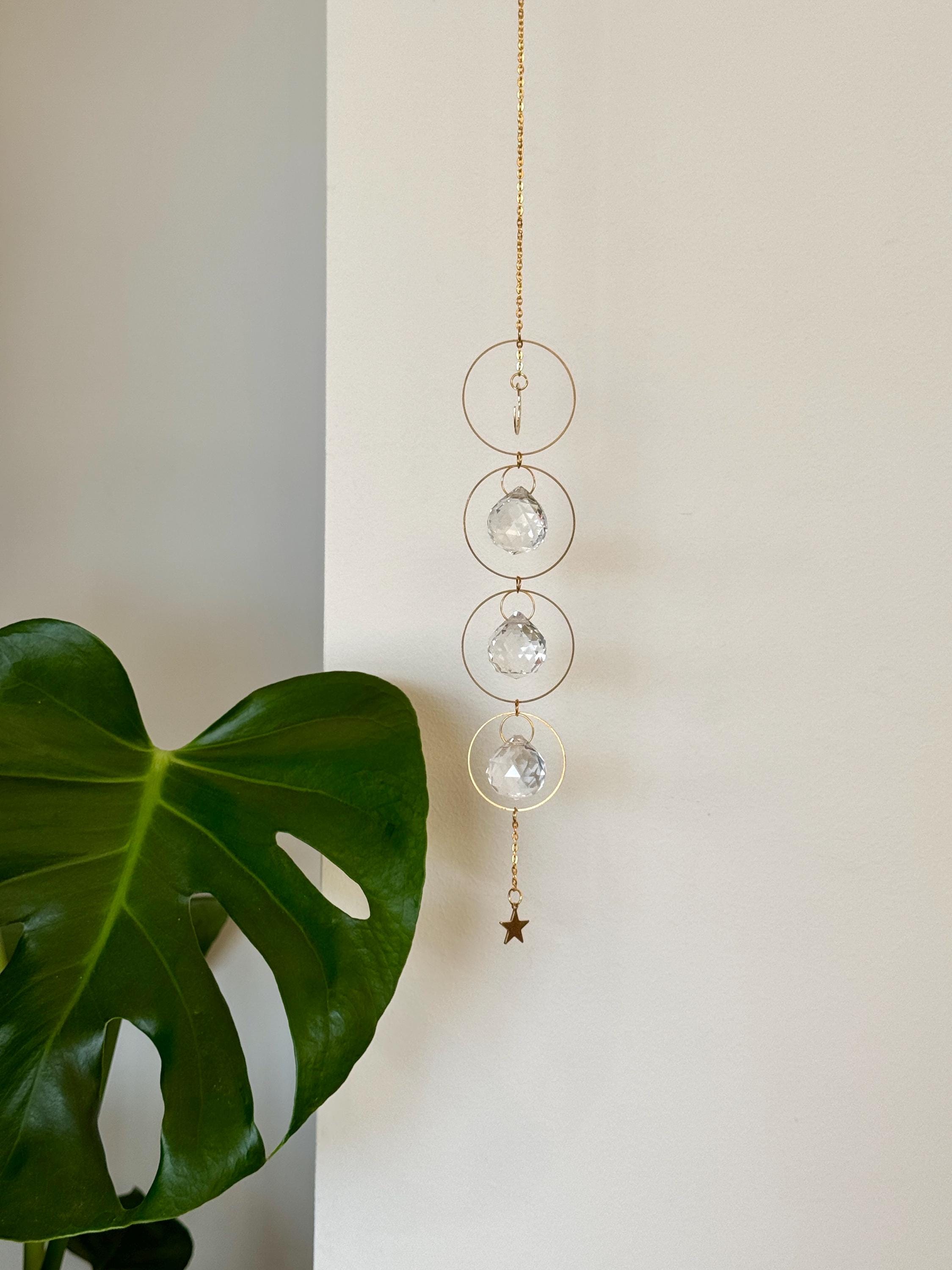 A gold-tone 'Serenity' celestial suncatcher hangs against a warm, neutral wall, featuring three circular brass hoops. The top hoop holds a delicate crescent moon, while the bottom three each frame a 20mm faceted crystal prism that ends in a small gold star. Bright sunlight creates a magical "Meer von Regenbögen" (sea of rainbows) and shimmering light speckles across the surface. To the right, a reed diffuser and stacked books create a sense of Gemütlichkeit (coziness), while the suncatcher casts a soft, rhy