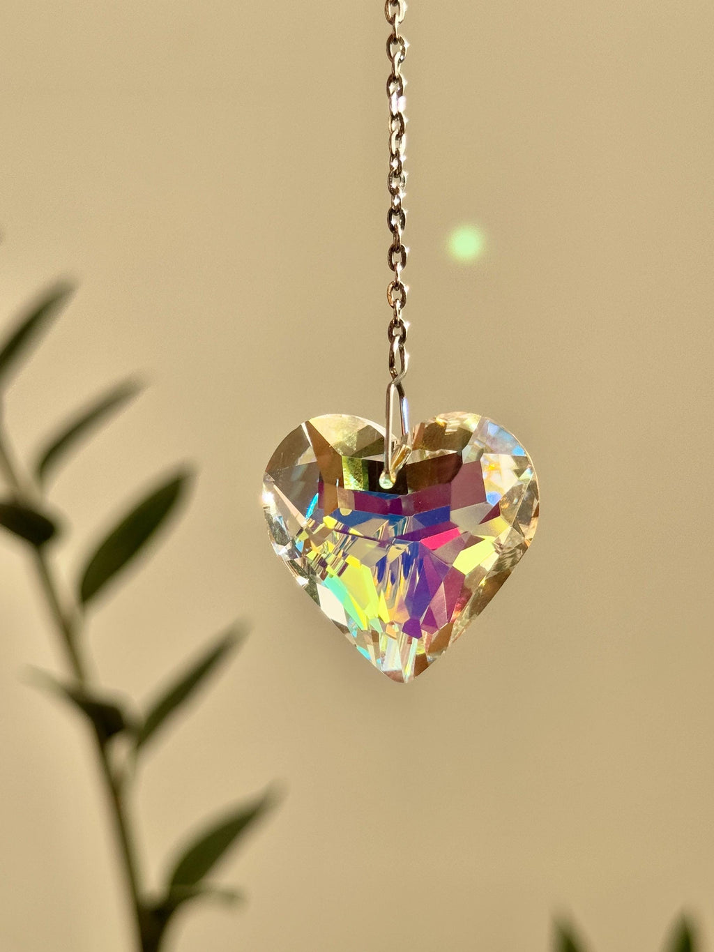 Close-up of 'Sweet Reflection' heart-shaped crystal suncatcher hanging on a silver stainless steel chain, casting vibrant rainbow prism patterns on a white wall next to a green Monstera leaf.
