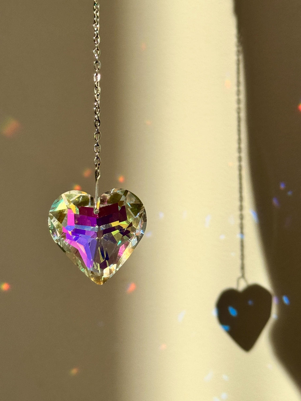 Close-up of 'Sweet Reflection' heart-shaped crystal suncatcher hanging on a silver stainless steel chain, casting vibrant rainbow prism patterns on a white wall next to a green Monstera leaf.
