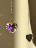 Close-up of 'Sweet Reflection' heart-shaped crystal suncatcher hanging on a silver stainless steel chain, casting vibrant rainbow prism patterns on a white wall next to a green Monstera leaf.