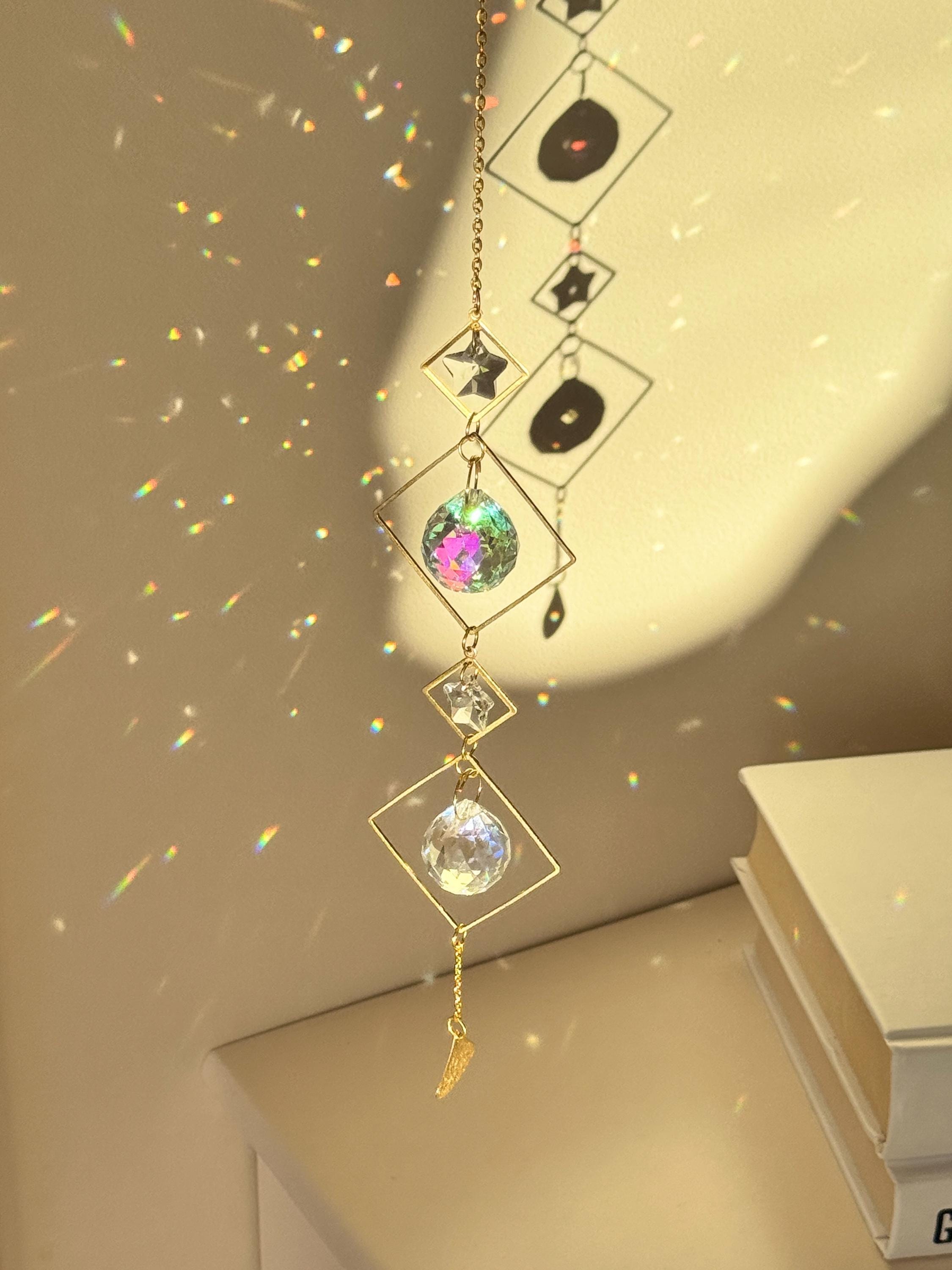 A vertical gold-tone geometric suncatcher hangs elegantly against a cream-colored wall, casting a dazzling "Meer von Regenbögen" (sea of rainbows) across the room. The piece features alternating small and large diamond-shaped brass frames, with two faceted 2cm crystal prisms suspended in the center. Sunlight passes through the glass, creating vibrant spectral flares that dance like "Sternenstaub" (stardust). To the right, a lush green Monstera leaf and a stack of books add a modern, boho-chic vibe. The over
