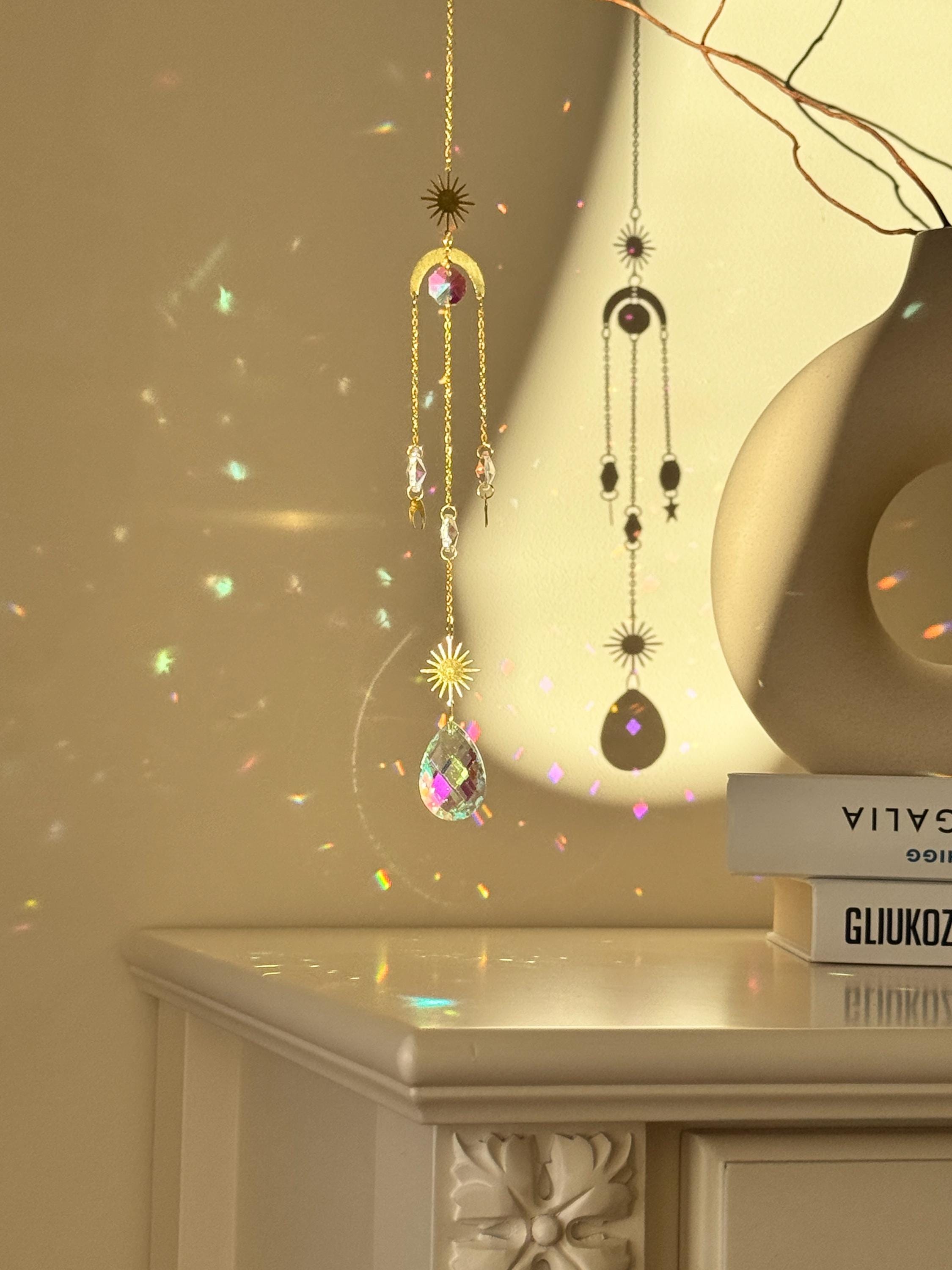 The 'Cosmic Balance' suncatcher, 47cm long, featuring a gold celestial geometric frame and a large 4cm crystal prism, hanging in a bright window and casting wide rainbow patterns.