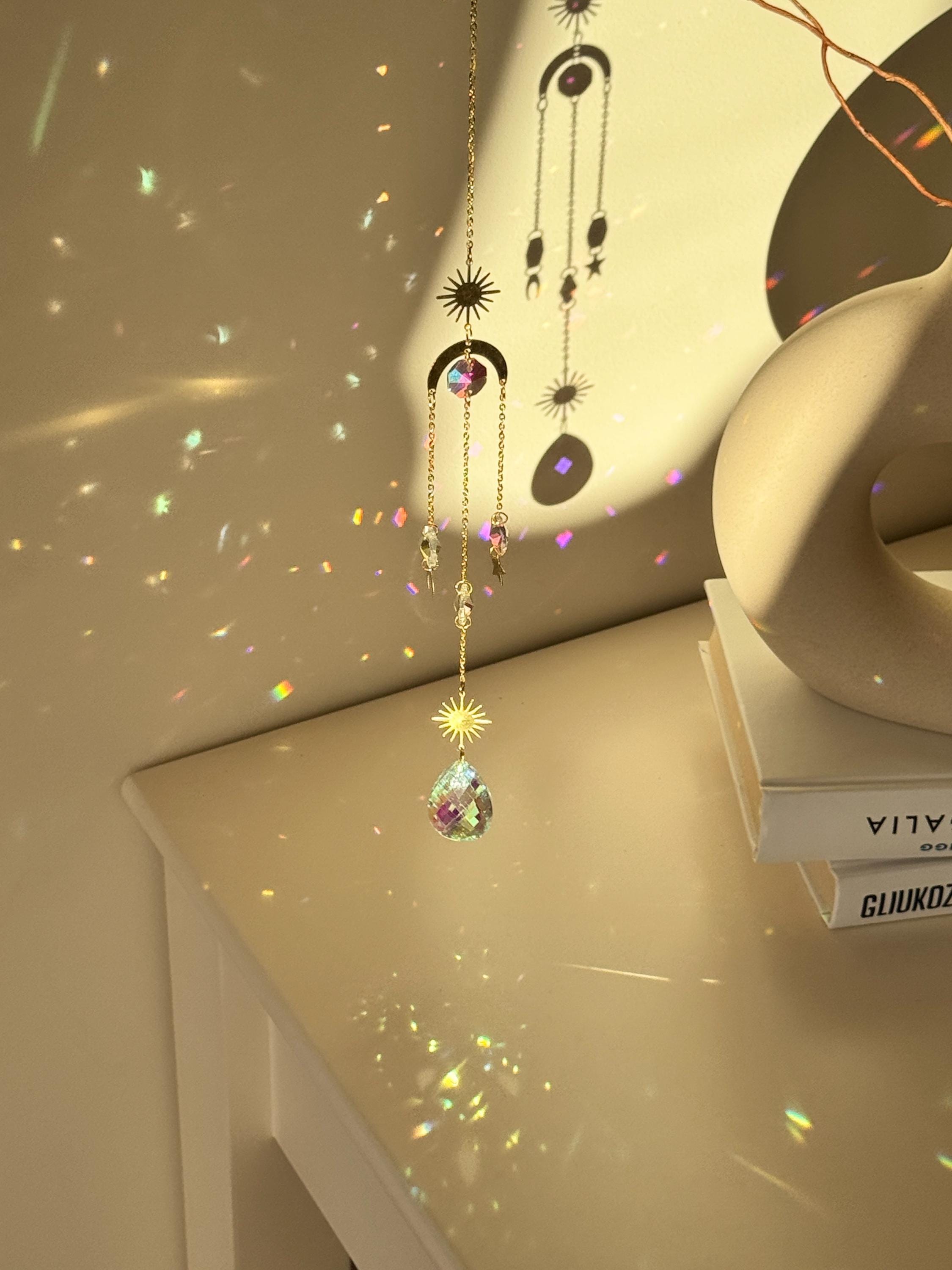 The 'Cosmic Balance' suncatcher, 47cm long, featuring a gold celestial geometric frame and a large 4cm crystal prism, hanging in a bright window and casting wide rainbow patterns.