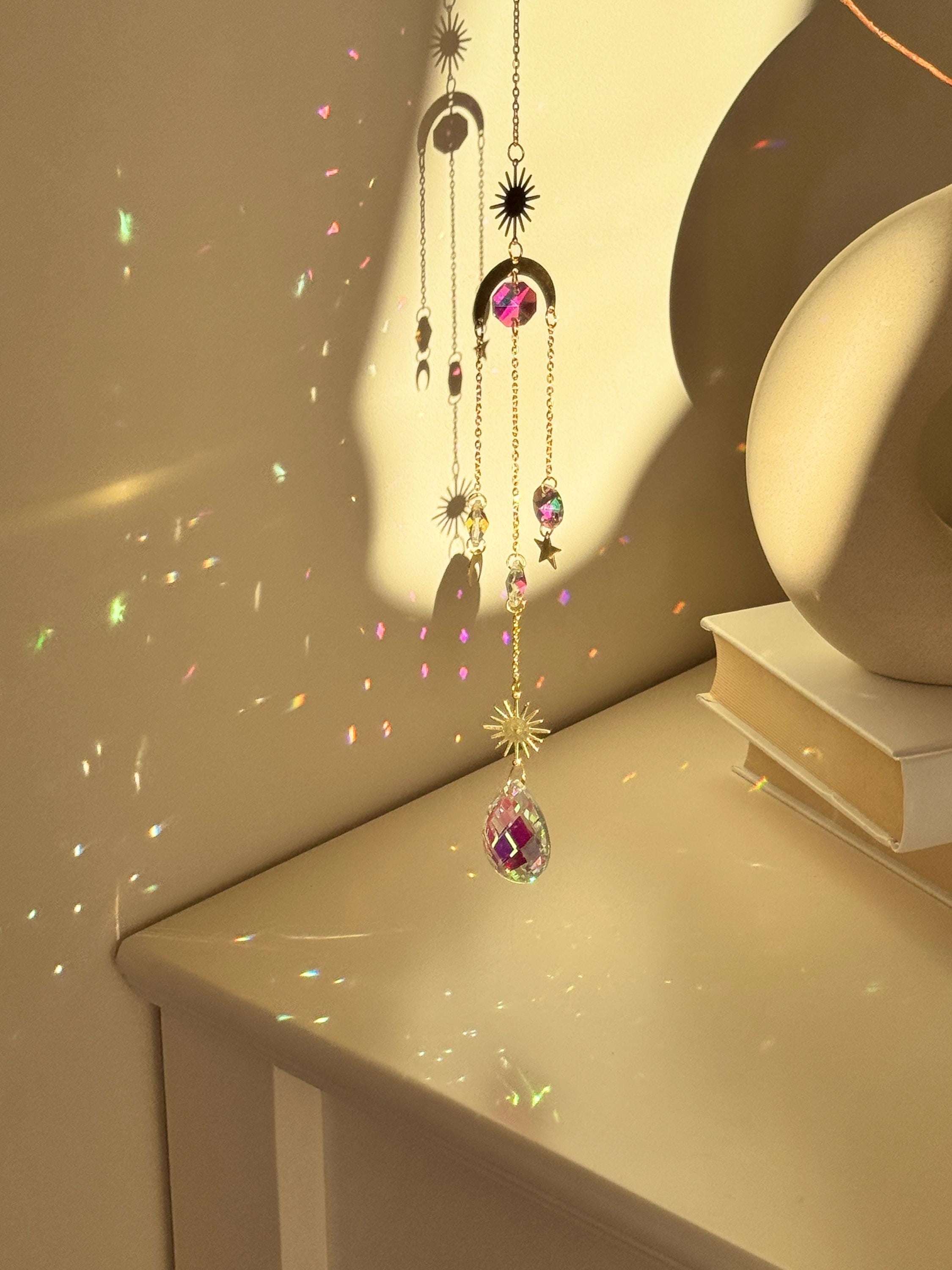 The 'Cosmic Balance' suncatcher, 47cm long, featuring a gold celestial geometric frame and a large 4cm crystal prism, hanging in a bright window and casting wide rainbow patterns.