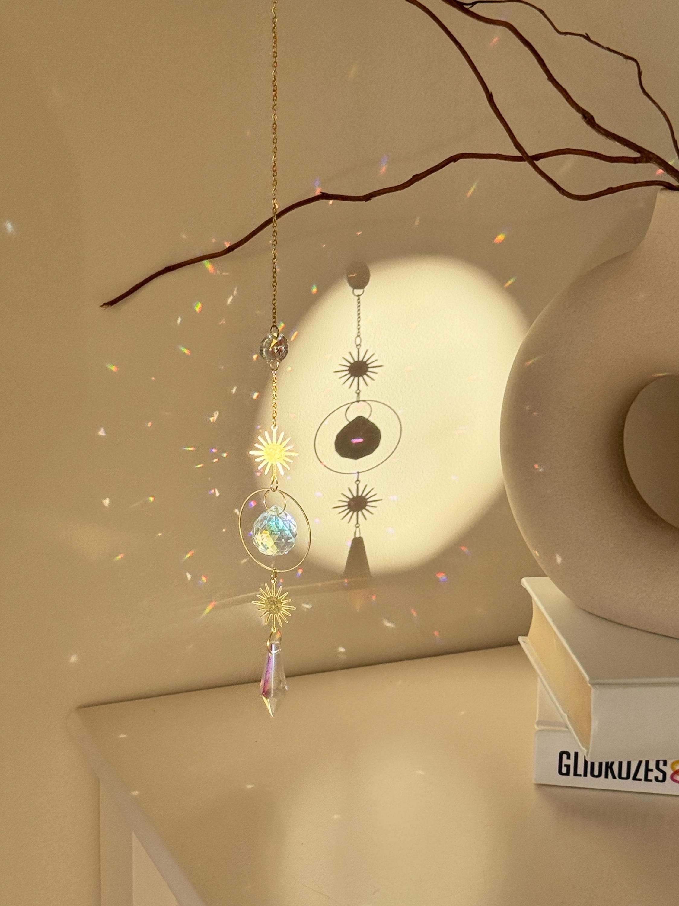 Celestial 'Sunny Days' suncatcher with gold sun charms and two crystal prisms hanging against a warm-toned wall, casting small rainbow light spots across the room.