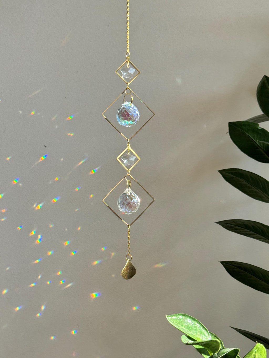A vertical gold-tone geometric suncatcher hangs elegantly against a cream-colored wall, casting a dazzling "Meer von Regenbögen" (sea of rainbows) across the room. The piece features alternating small and large diamond-shaped brass frames, with two faceted 2cm crystal prisms suspended in the center. Sunlight passes through the glass, creating vibrant spectral flares that dance like "Sternenstaub" (stardust). To the right, a lush green Monstera leaf and a stack of books add a modern, boho-chic vibe. The over