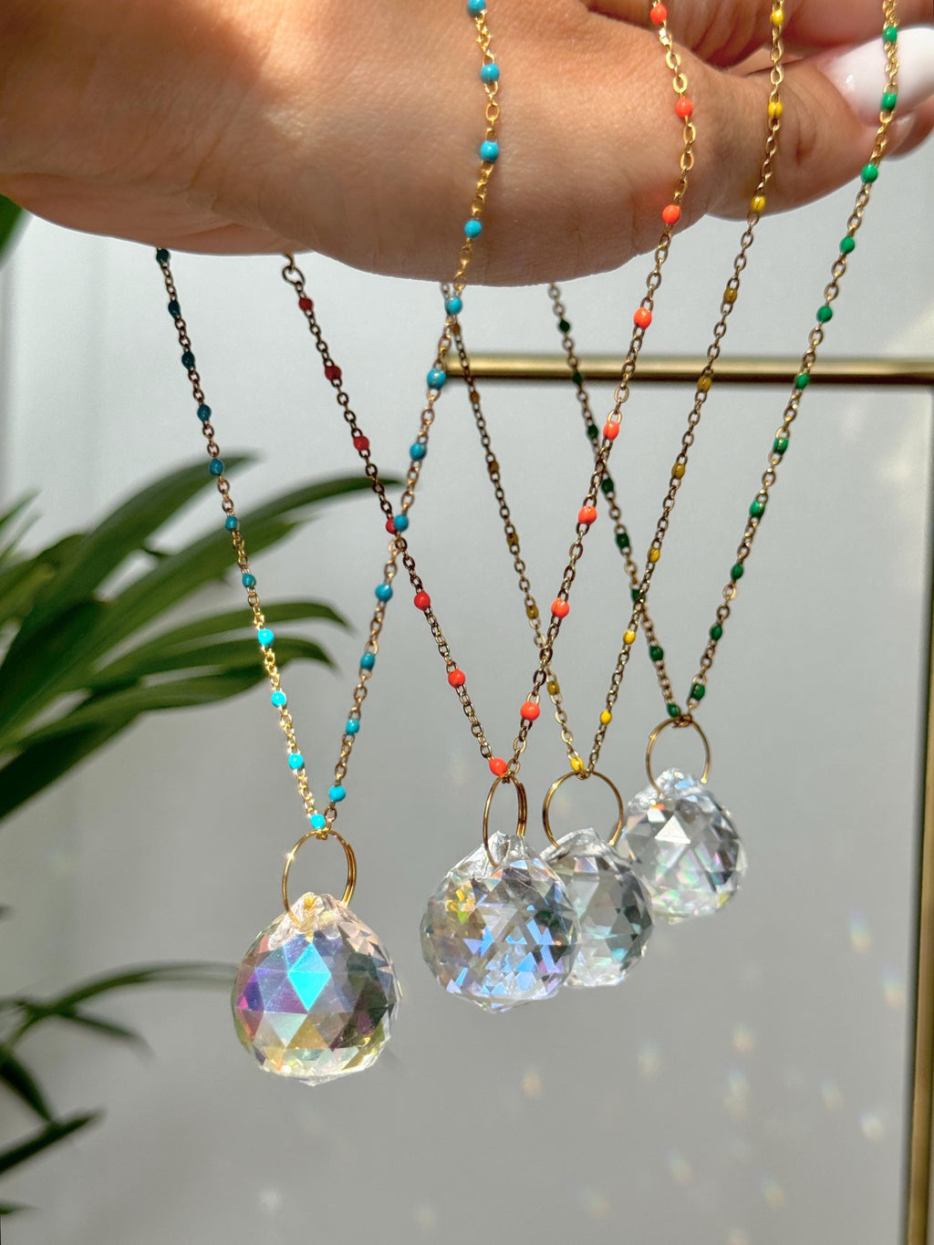 Four crystal prism car suncatchers in different sizes hanging from a hand, casting rainbow light reflections on a white wall next to a green plant.
