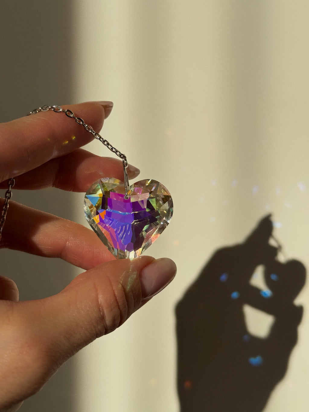 Close-up of 'Sweet Reflection' heart-shaped crystal suncatcher hanging on a silver stainless steel chain, casting vibrant rainbow prism patterns on a white wall next to a green Monstera leaf.