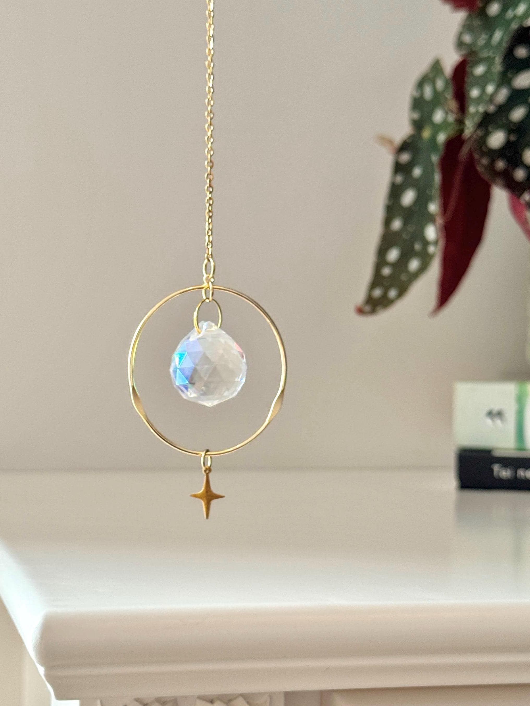 Celestial Compass suncatcher (Sonnenfänger) with a gold wavy brass ring,  crystal prism, and star charm, reflecting rainbows (Regenbogen) on a white wall.