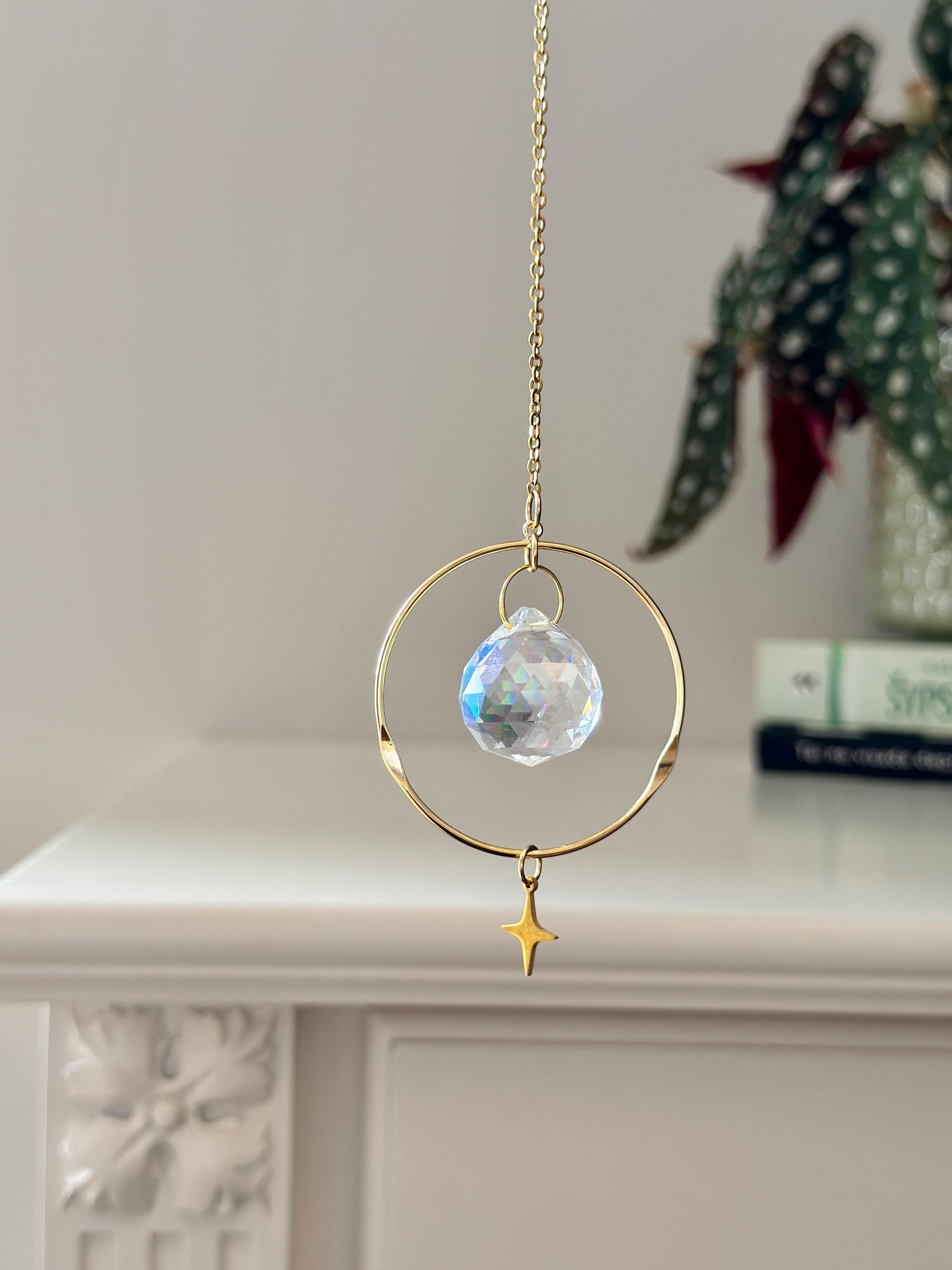 Celestial Compass suncatcher (Sonnenfänger) with a gold wavy brass ring,  crystal prism, and star charm, reflecting rainbows (Regenbogen) on a white wall.