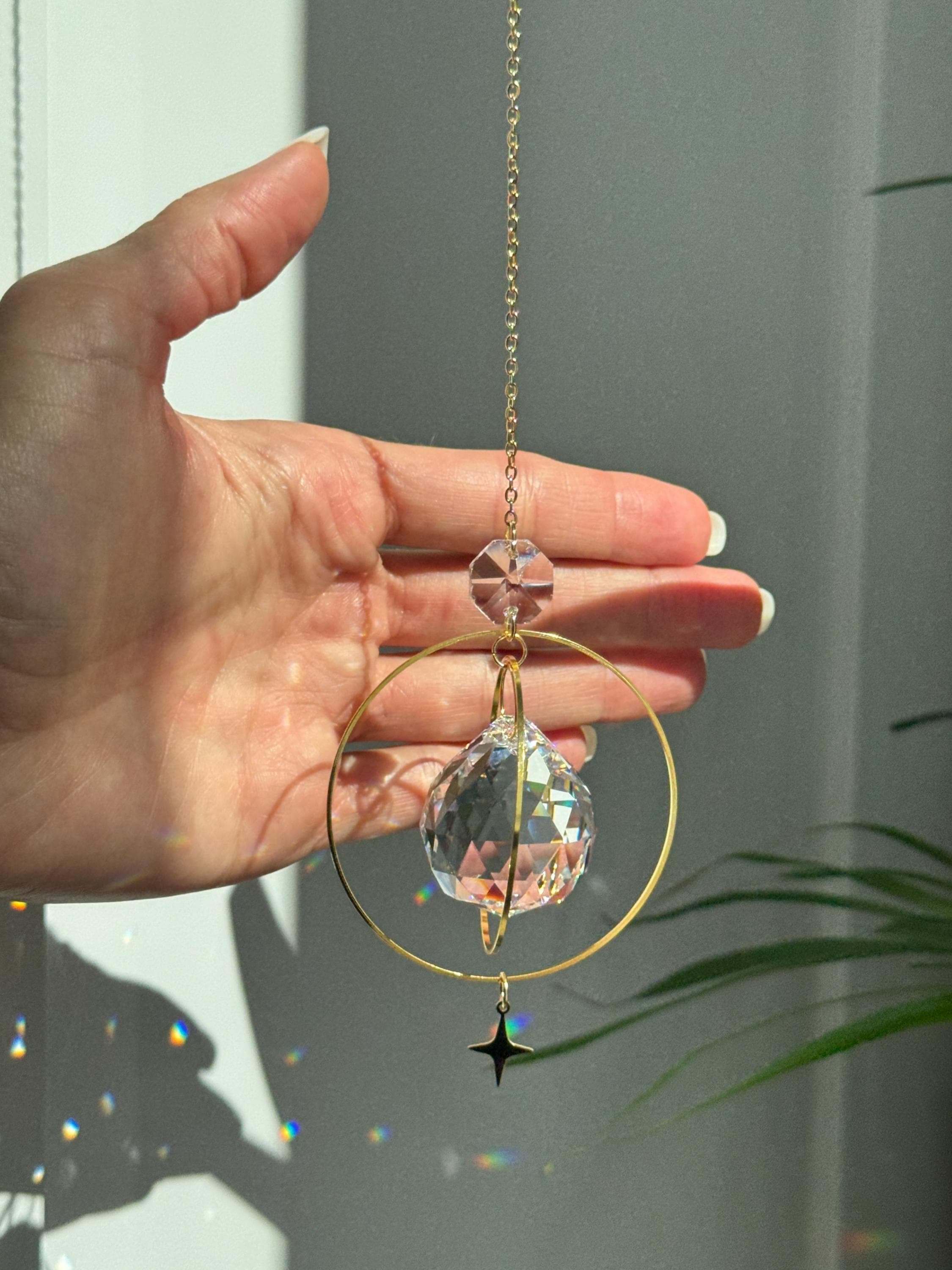The "North Star Prism" celestial suncatcher with a clear, high-quality faceted round crystal sphere. It features a smaller octagonal crystal bead and a sharp four-pointed gold star charm hanging below, reflecting brilliant light patterns on the wall behind.