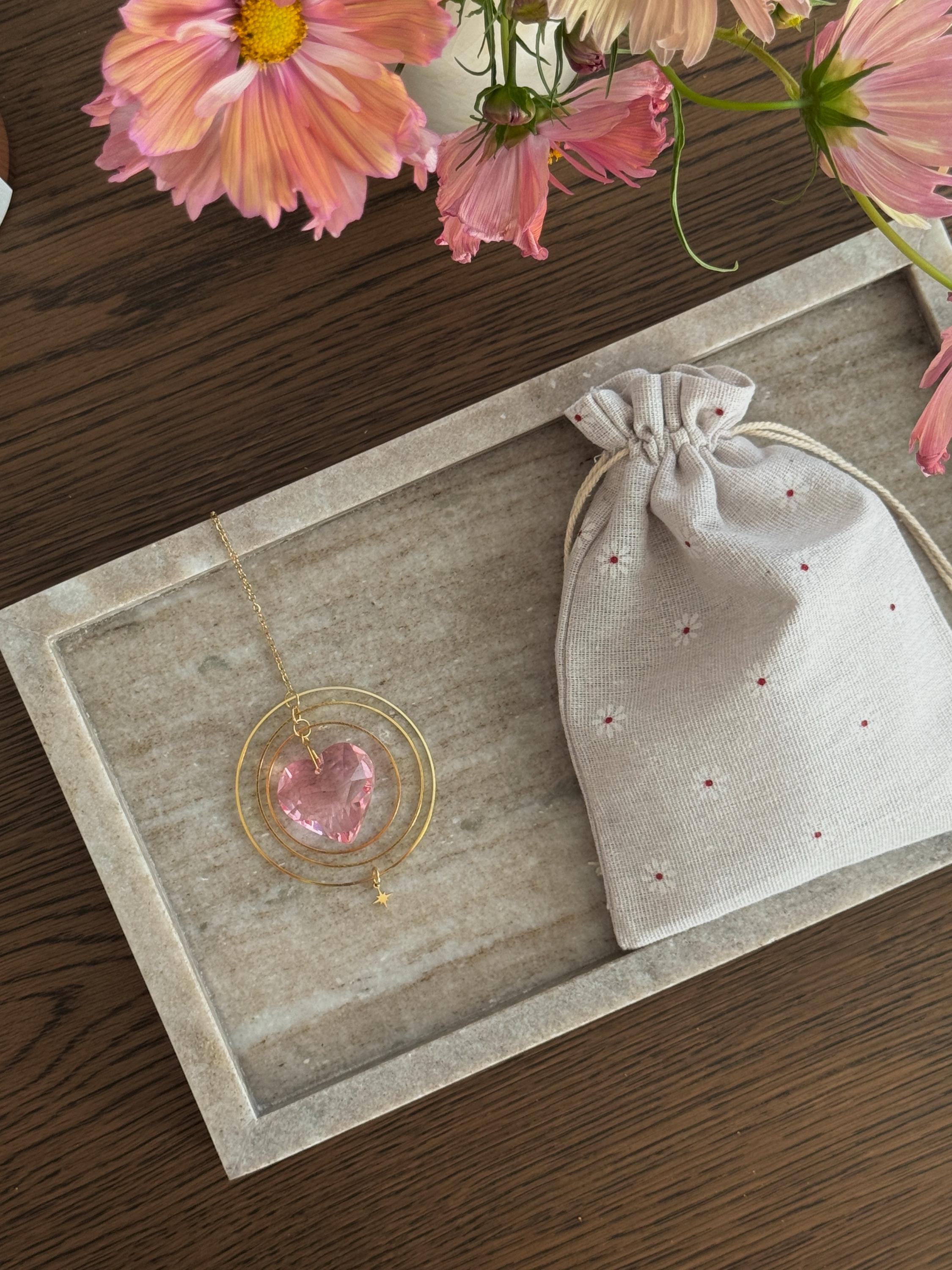 heart shaped suncatcher on a stone tray with a linen drawstring bag and pink flowers in the background