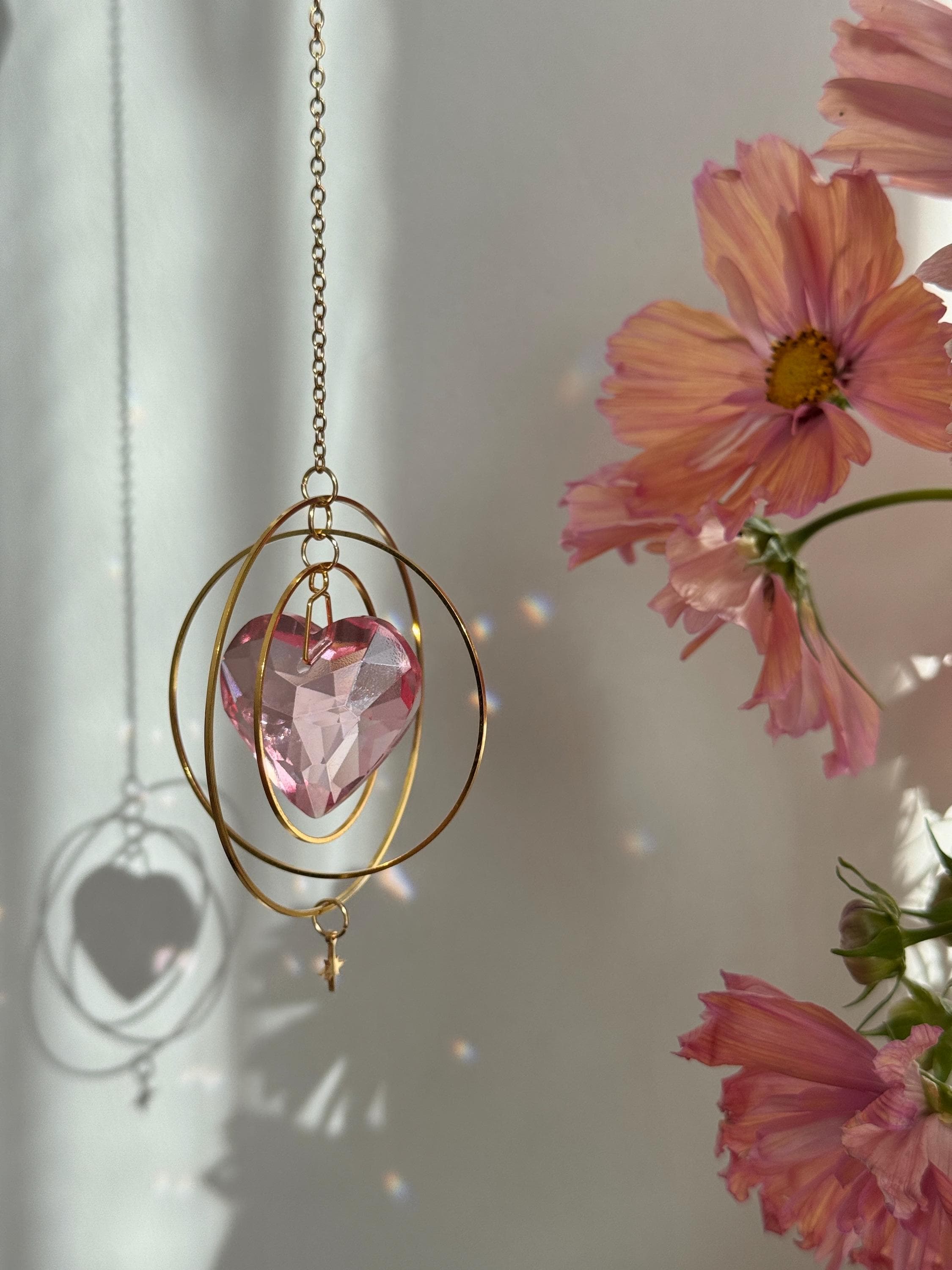 Sunkissed Heartbeat crystal suncatcher featuring a pink-gold faceted heart prism. Handmade celestial window decor with gold rings casting rainbow light flares (Sonnenfänger).