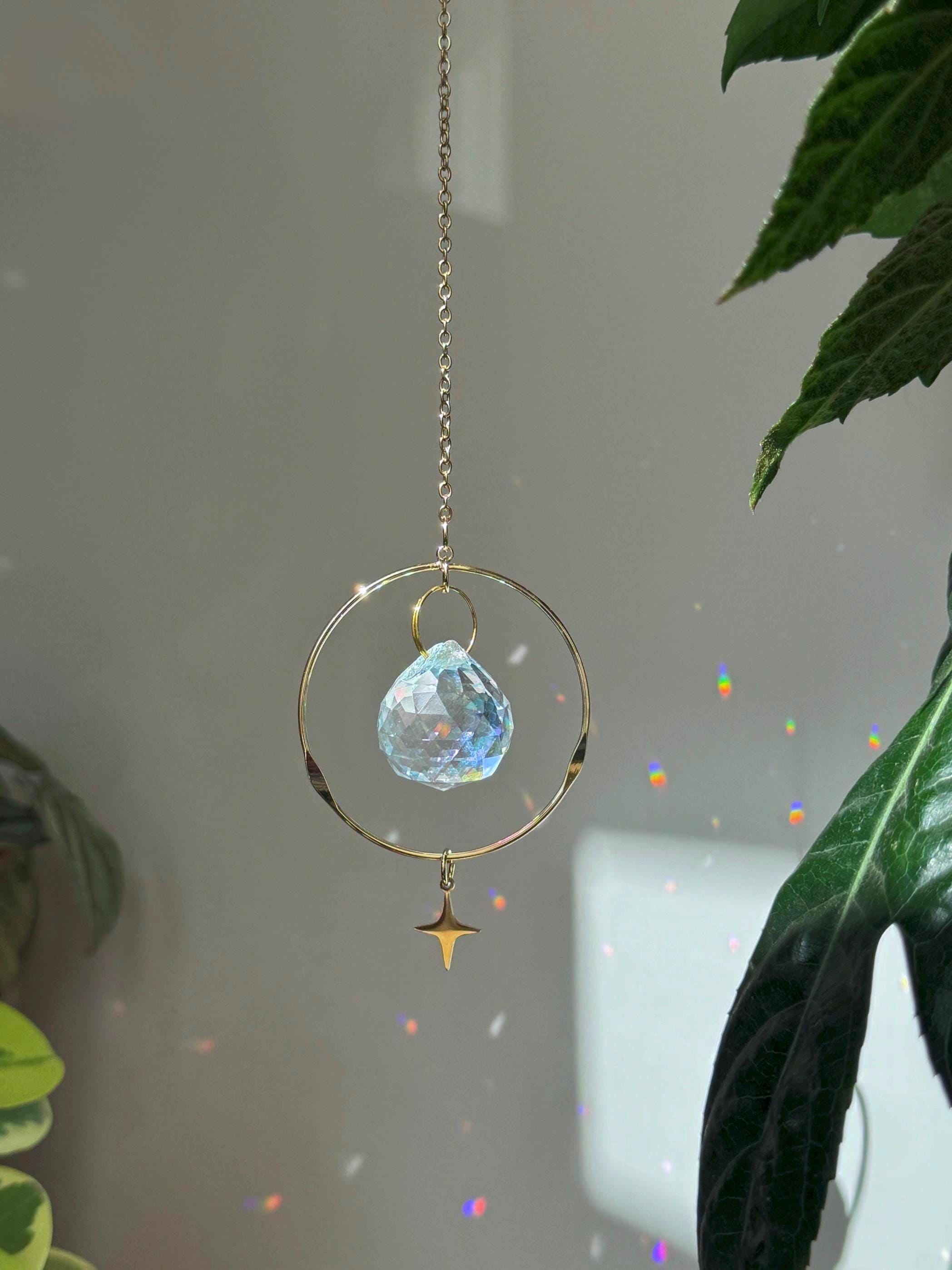 Celestial Compass suncatcher (Sonnenfänger) with a gold wavy brass ring,  crystal prism, and star charm, reflecting rainbows (Regenbogen) on a white wall.