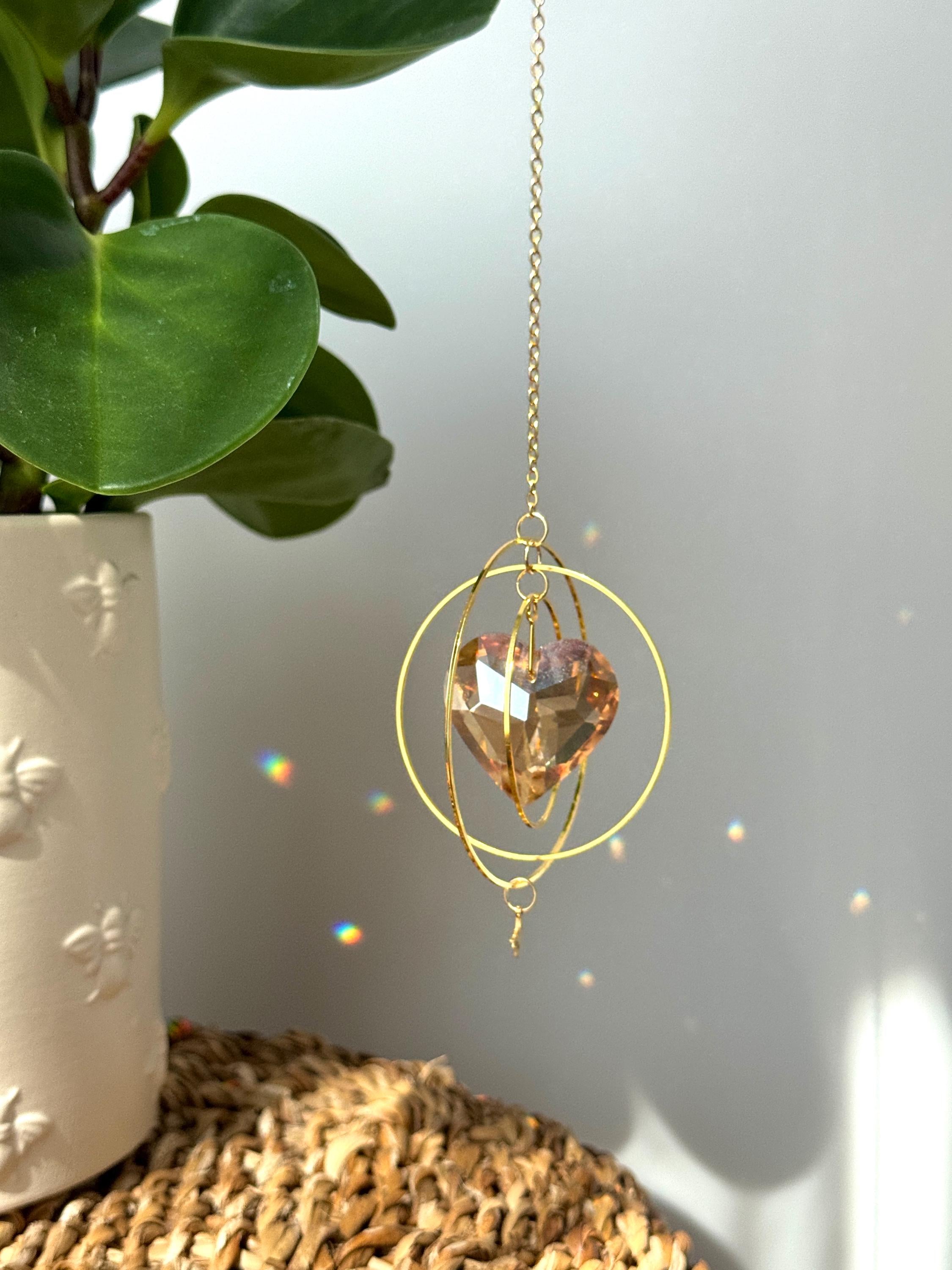Sunkissed Heartbeat crystal suncatcher featuring a warm amber-gold faceted heart prism. Handmade celestial window decor with gold rings casting rainbow light flares (Sonnenfänger).