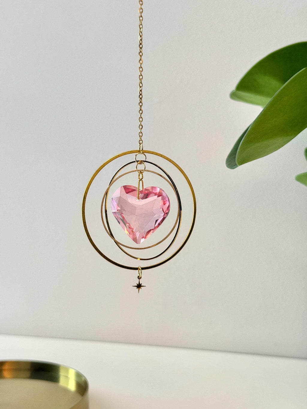 Sunkissed Heartbeat crystal suncatcher featuring a pink-gold faceted heart prism. Handmade celestial window decor with gold rings casting rainbow light flares (Sonnenfänger).