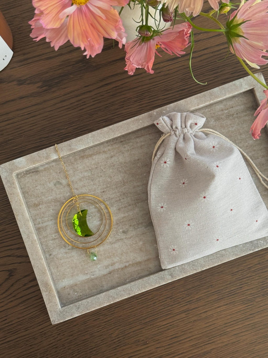 Over the Moon crystal suncatcher with elegant linen gift pouch. Green crescent moon prism on a marble tray next to pink flowers—perfect celestial gift idea (Geschenkidee).